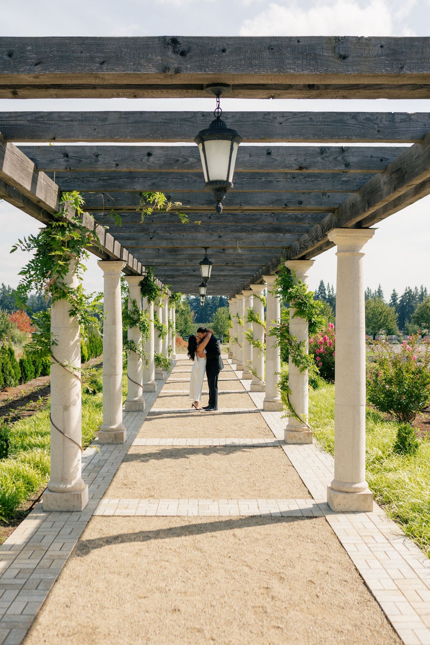 Long covered outdoor walkway with white columns and wooden ceiling leads through a garden landscape at dusk.