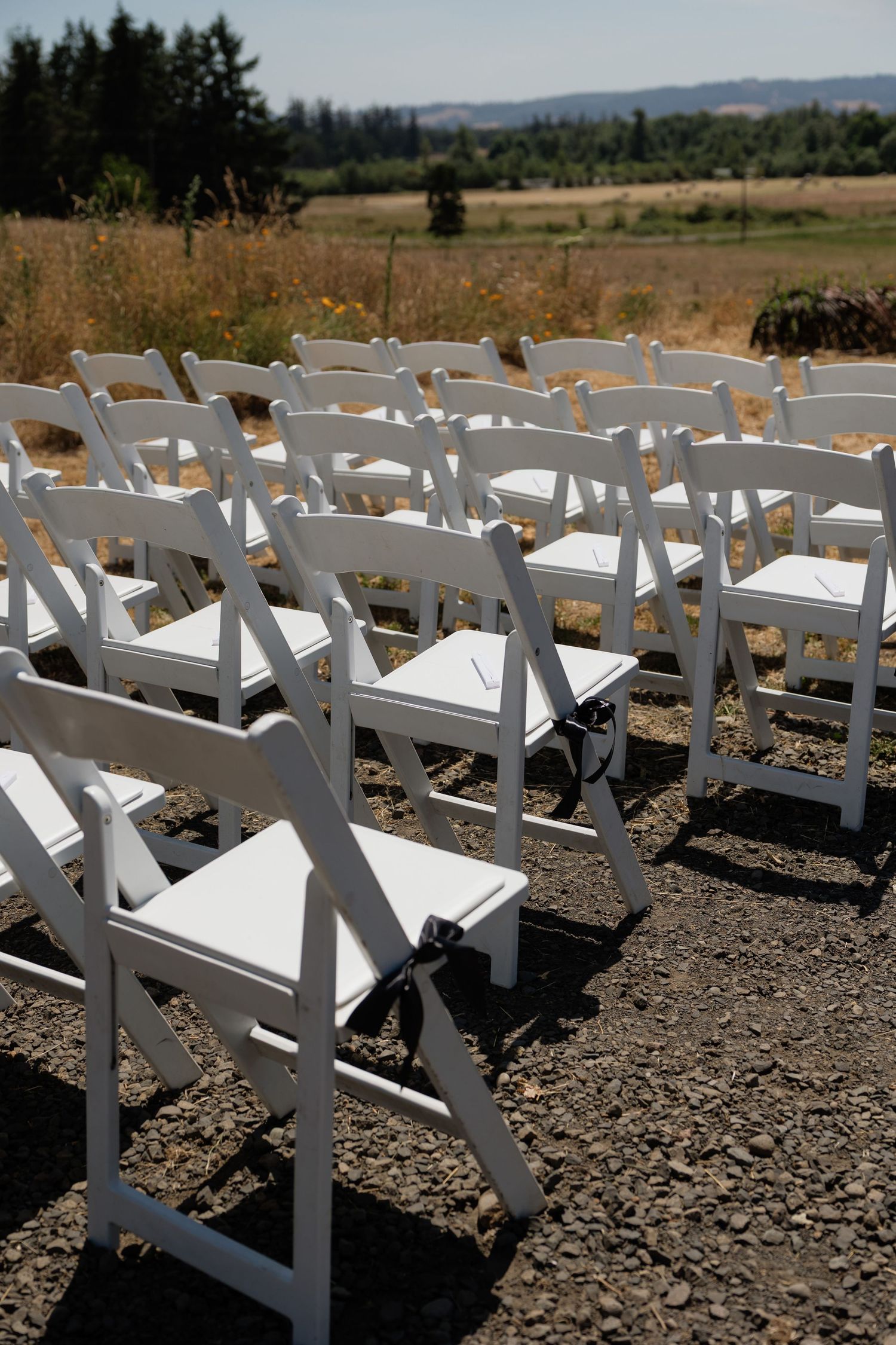Rows of white folding chairs arranged on gravel for an outdoor wedding ceremony with scenic field backdrop.