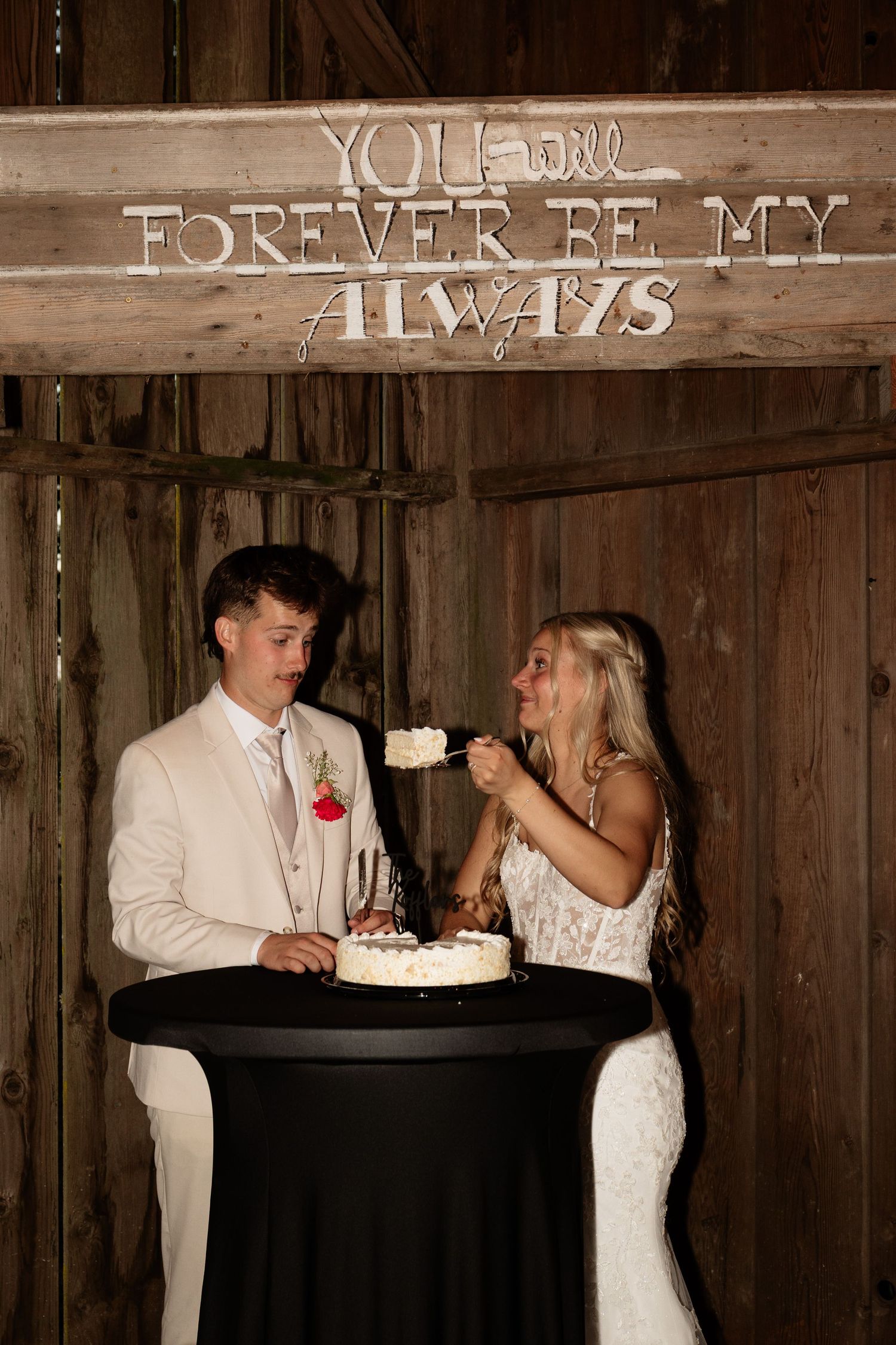Bride and groom cutting wedding cake under rustic wooden sign reading You Will Forever Be My Always.
