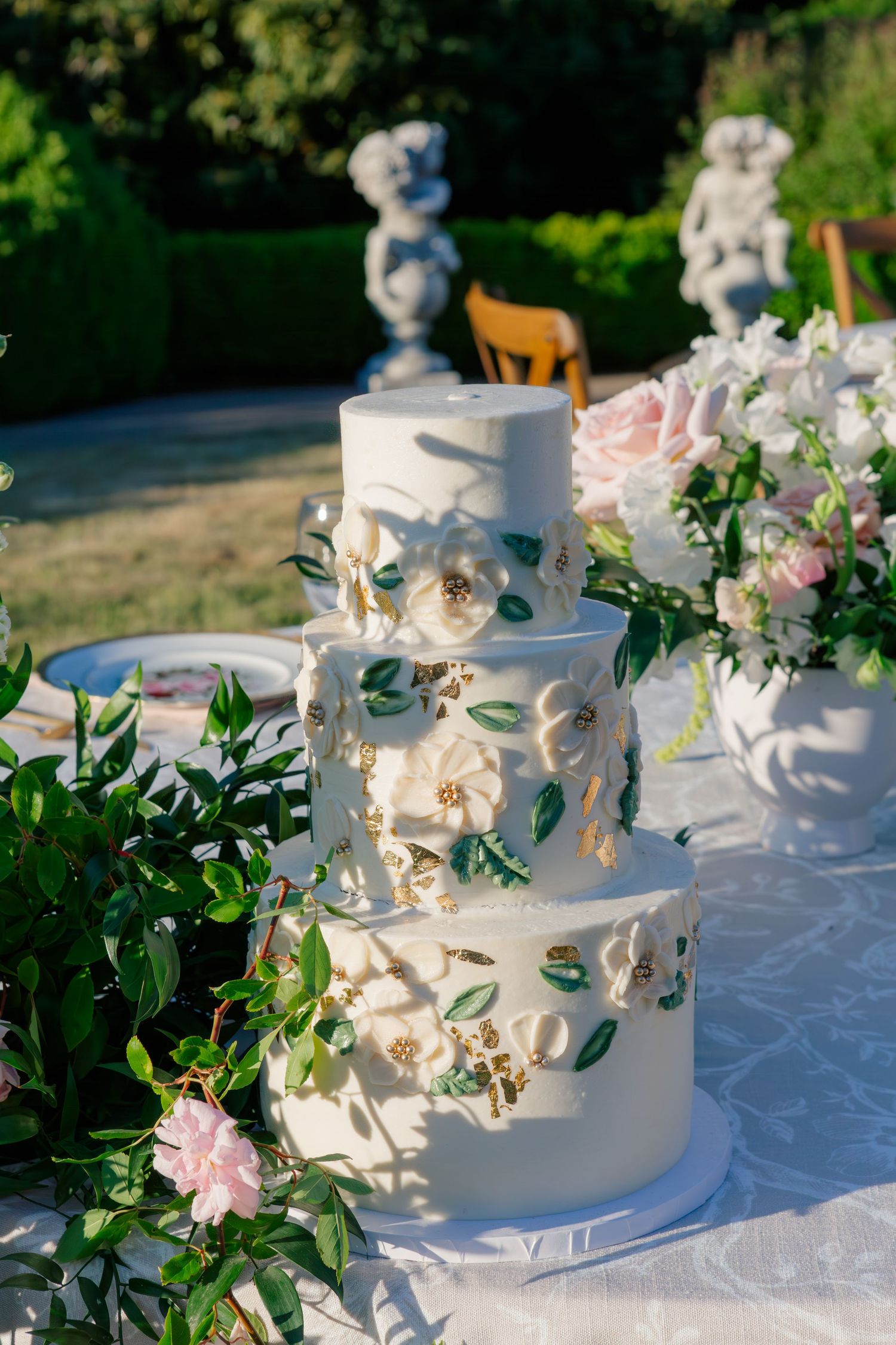 Elegant white wedding cake adorned with pink flowers and green foliage sits on linen-covered table in outdoor setting.