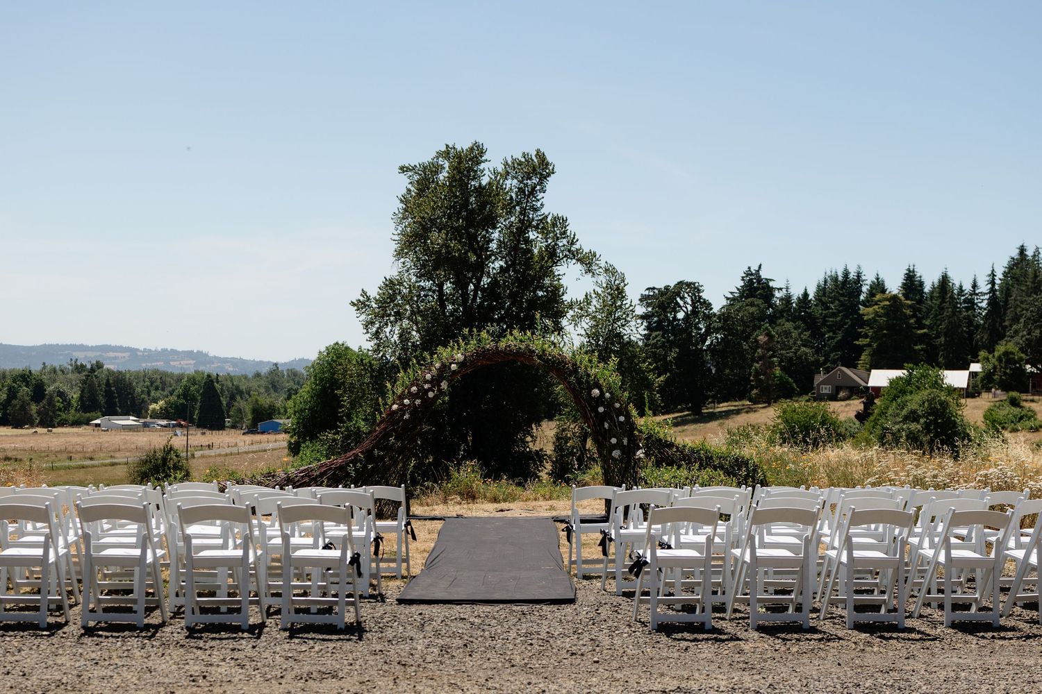 Outdoor wedding ceremony setup with white chairs, floral arch, and scenic countryside views under a clear blue sky.