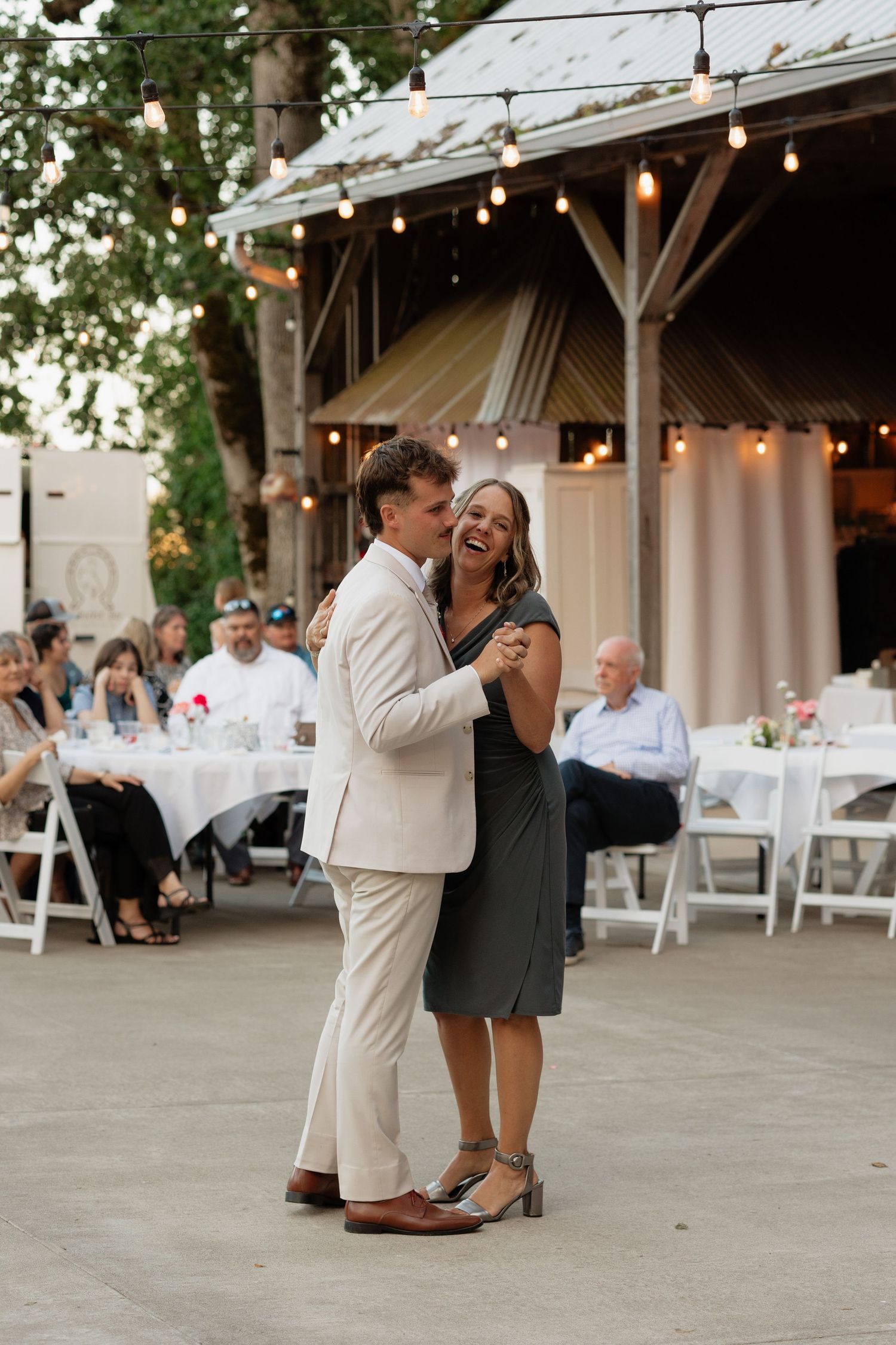 Groom in tan suit shares first dance with mother in green dress at outdoor wedding reception.