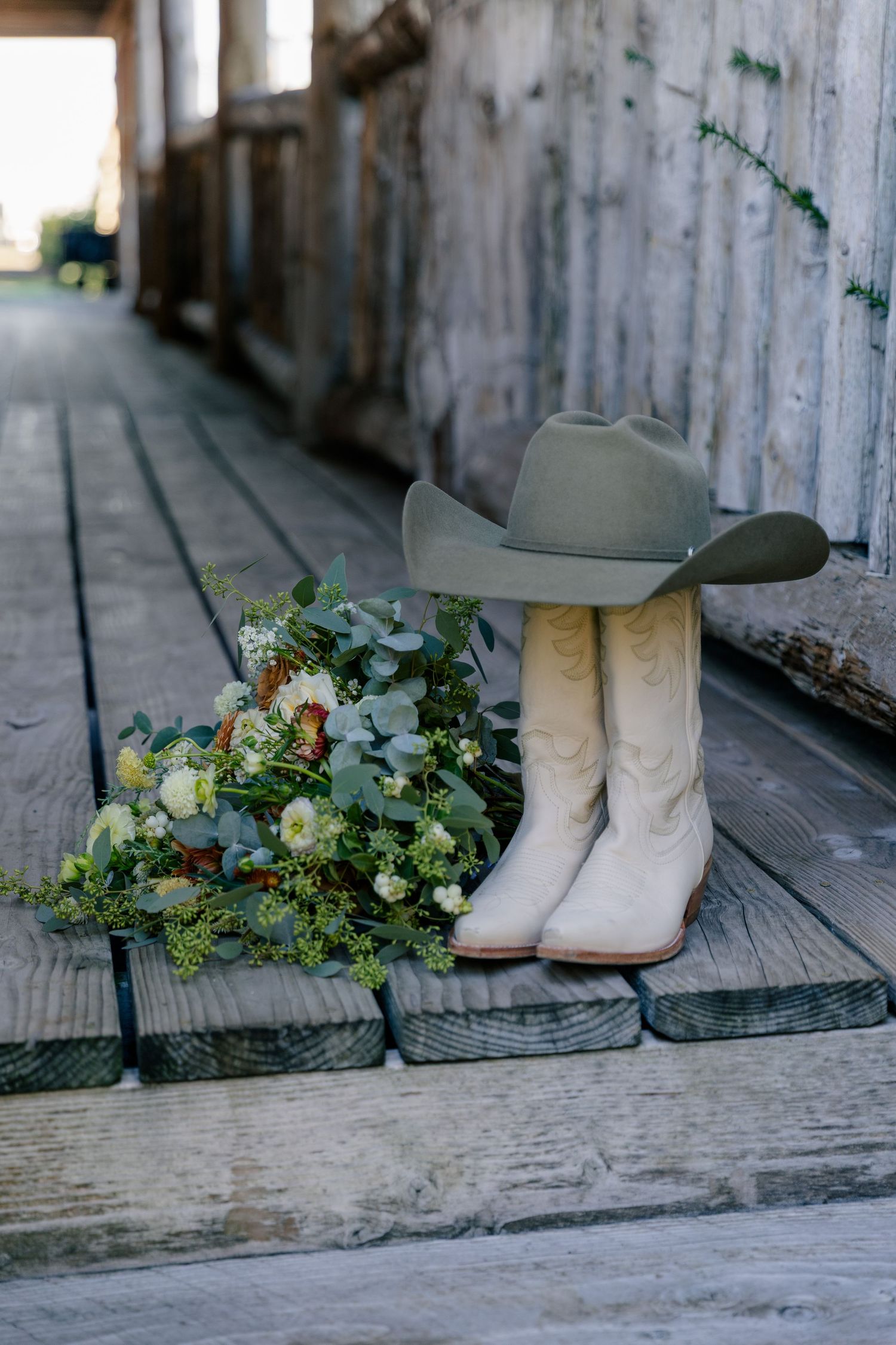 A rustic wedding detail shot showing white cowboy boots and a floral bouquet arranged on weathered wooden boards.