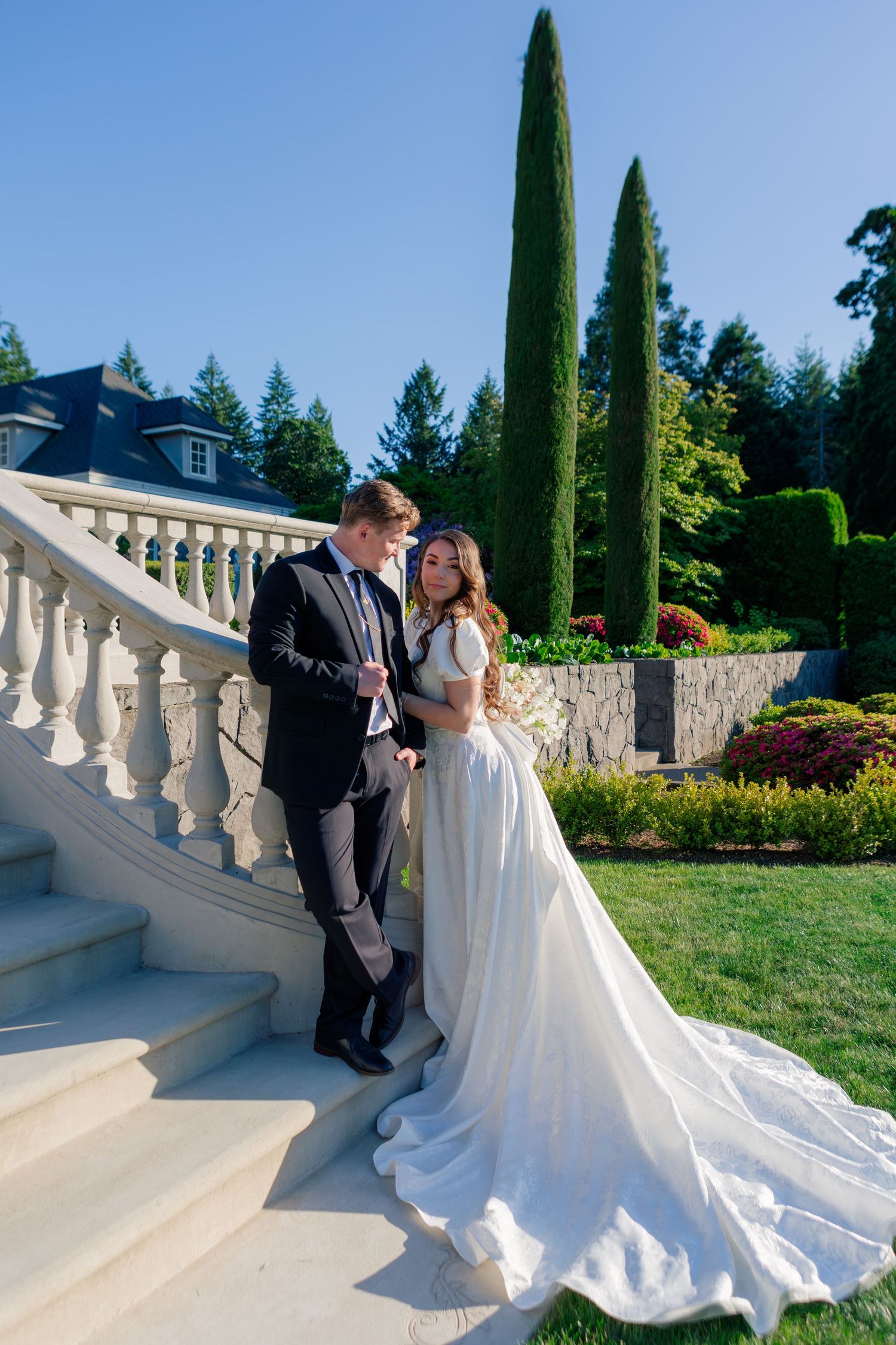 A couple in wedding attire pose on grand stone staircase with ornate railing and tall cypress trees in elegant garden setting.