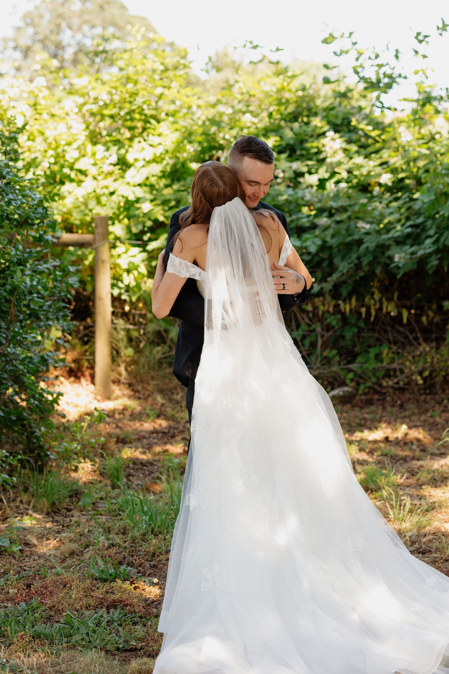 Bride and groom embracing in garden, wedding dress with veil trailing.