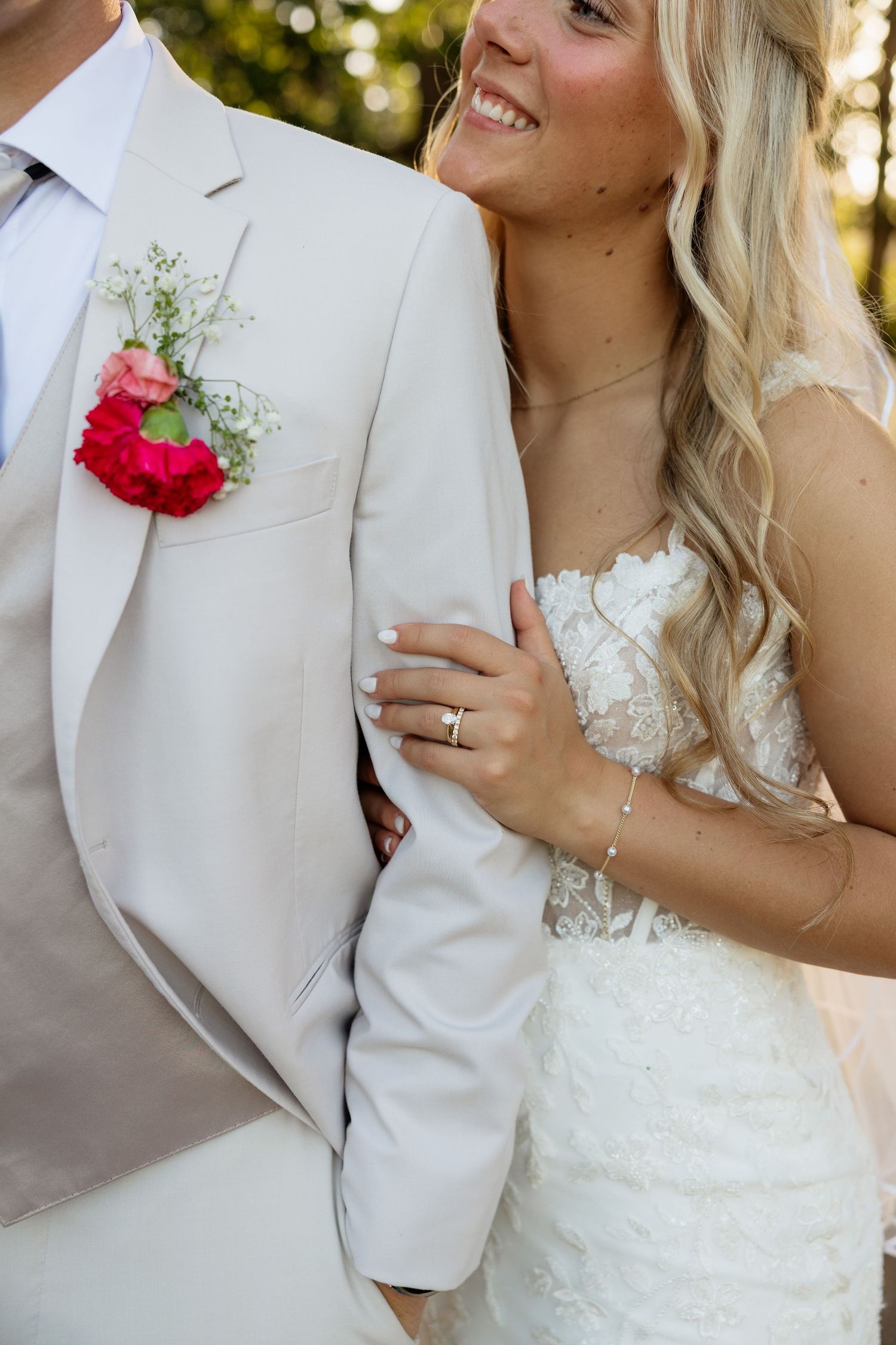 Bride in lace gown holding grooms beige suit jacket arm with wedding ring visible, smiling.