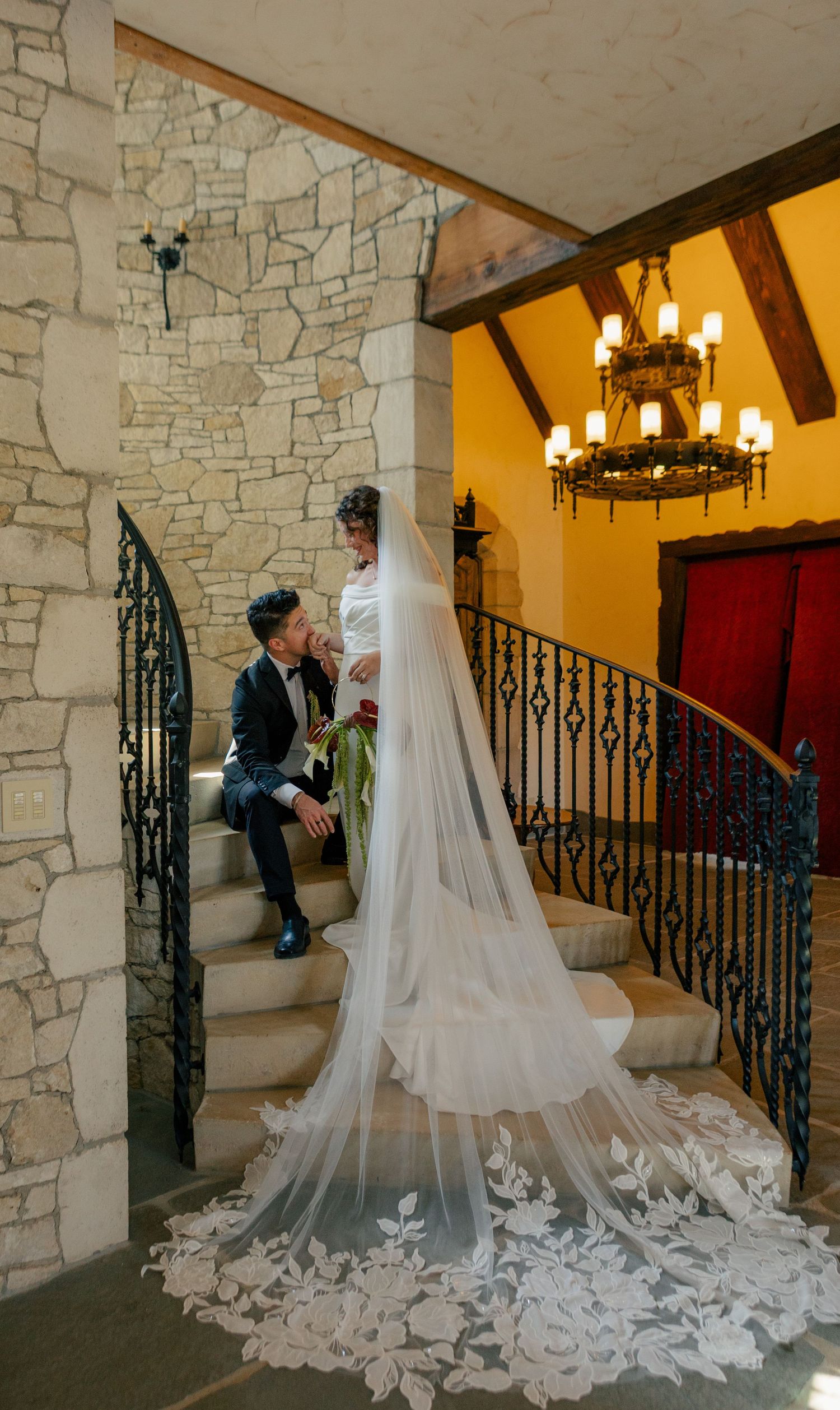 A bride in a flowing cathedral veil with floral details stands on stone steps near wrought iron railings in warm lighting.