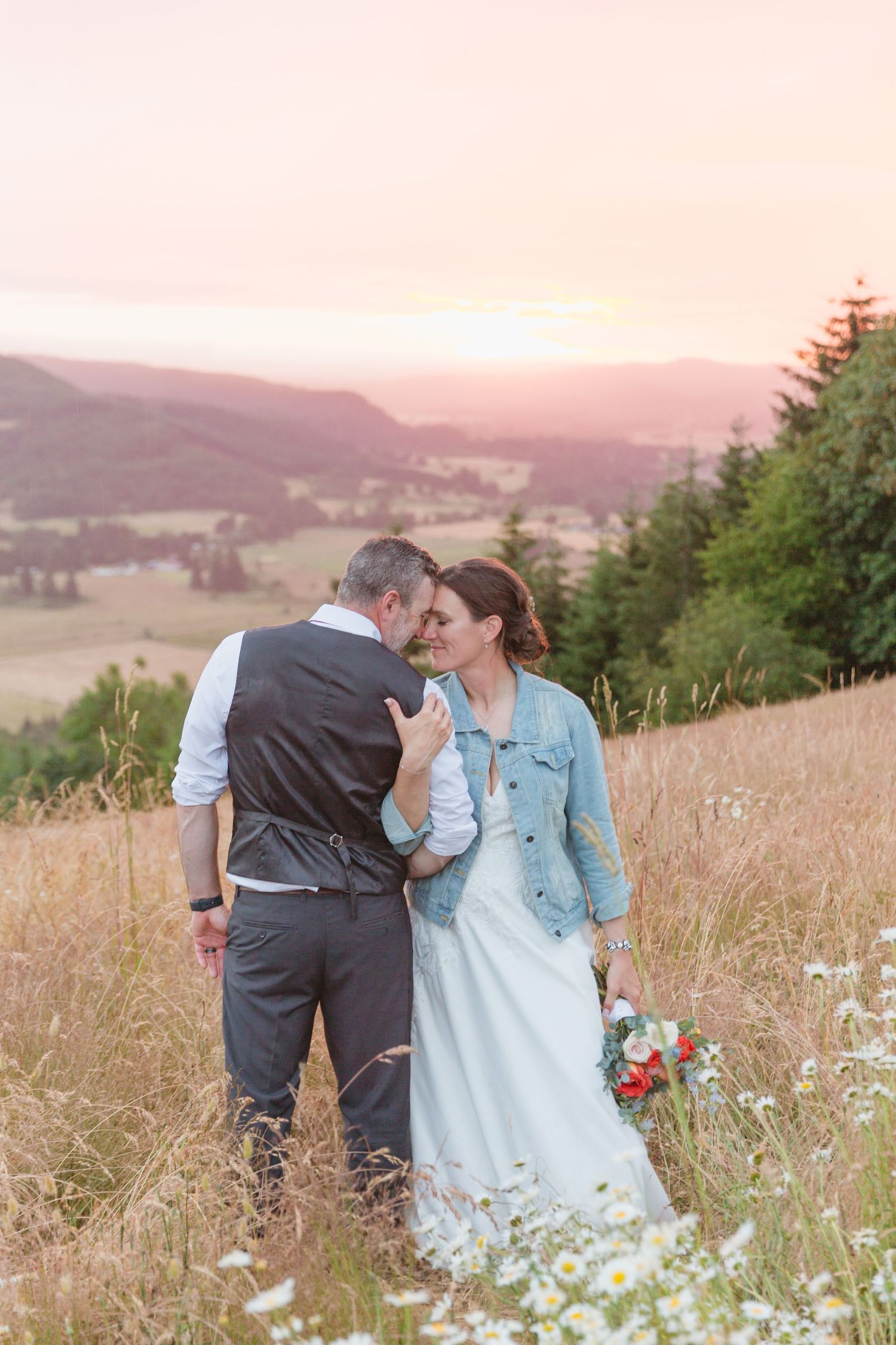 A couple embraces on a hillside at sunset with mountain views and wildflowers in a romantic outdoor setting.