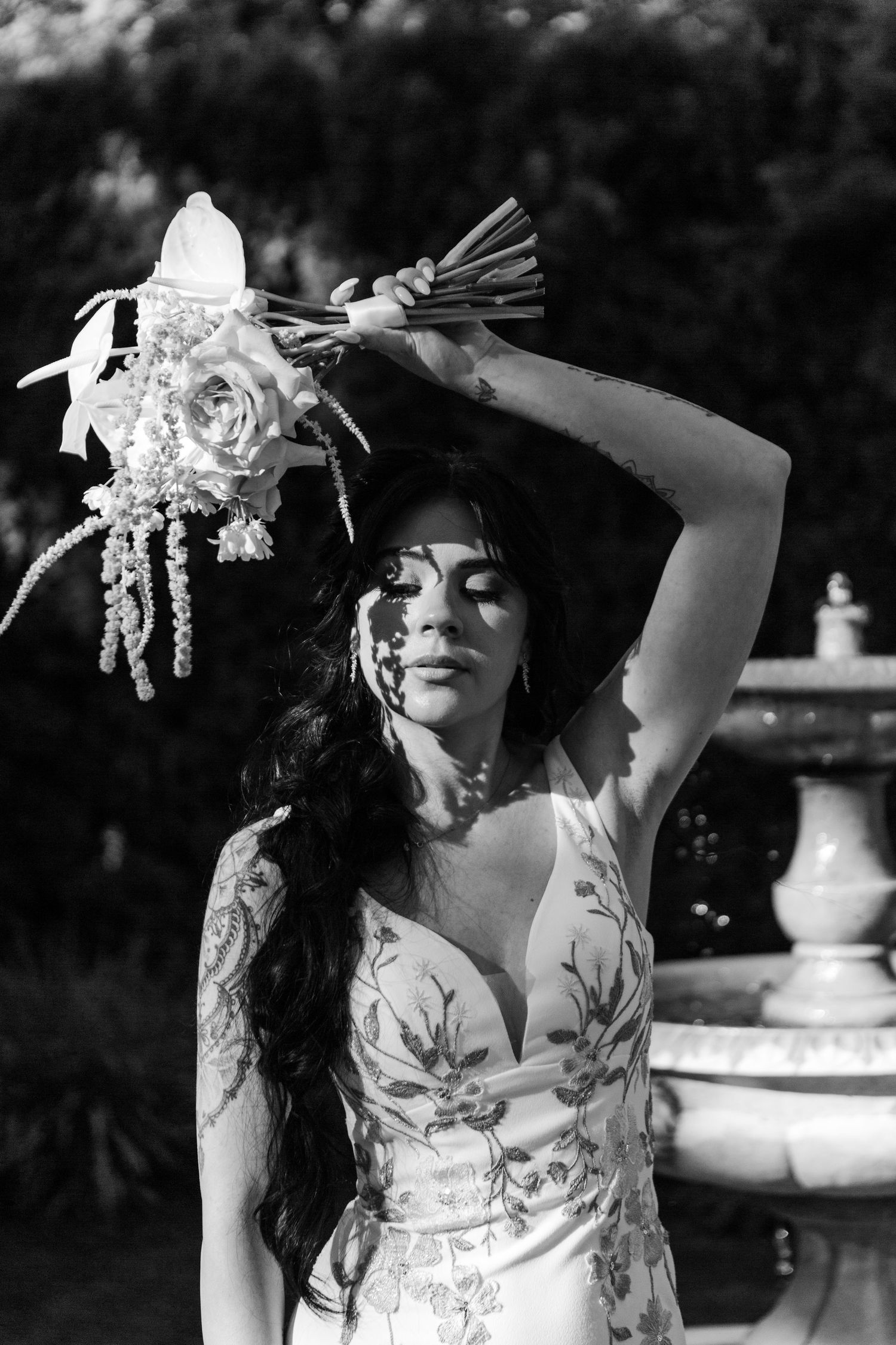Black and white photo of a bride in floral dress holding up her bouquet in an outdoor garden setting.