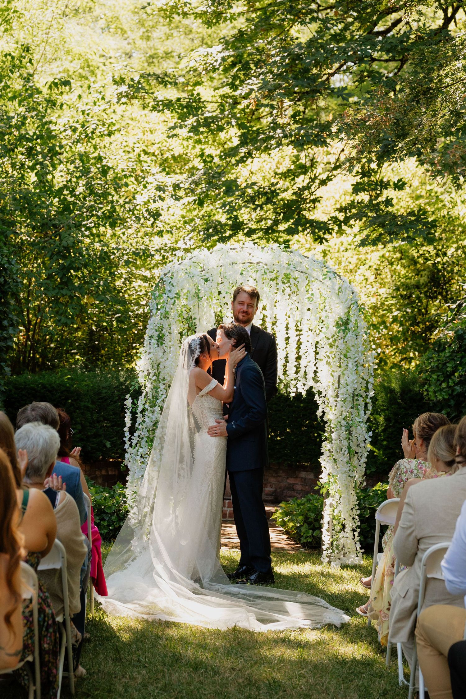 Bride and groom share first kiss under floral arch adorned with white wisteria during outdoor garden wedding ceremony.