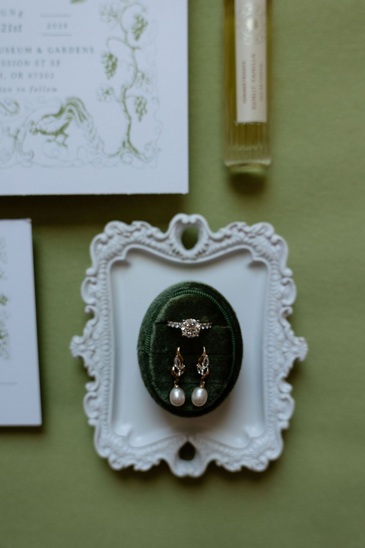 Pearl drop earrings and diamond ring in green velvet box on ornate white frame with wedding stationery.
