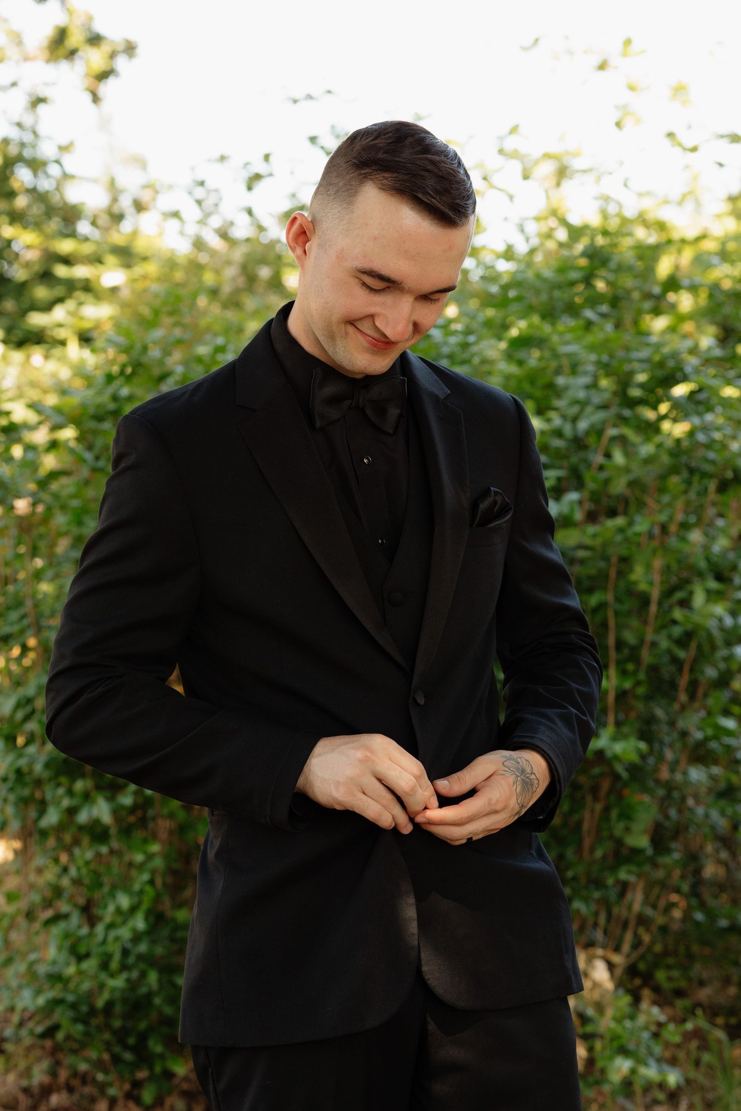 Elegant groom in black tuxedo buttoning jacket while smiling outdoors among green foliage.