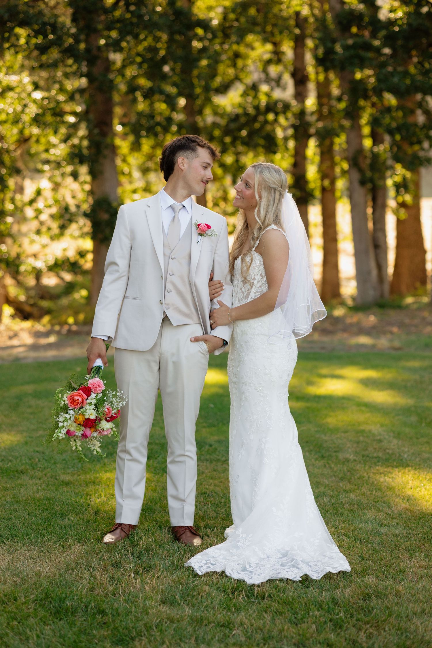 Newlywed couple in beige suit and lace gown share romantic glance on sunlit grassy lawn.