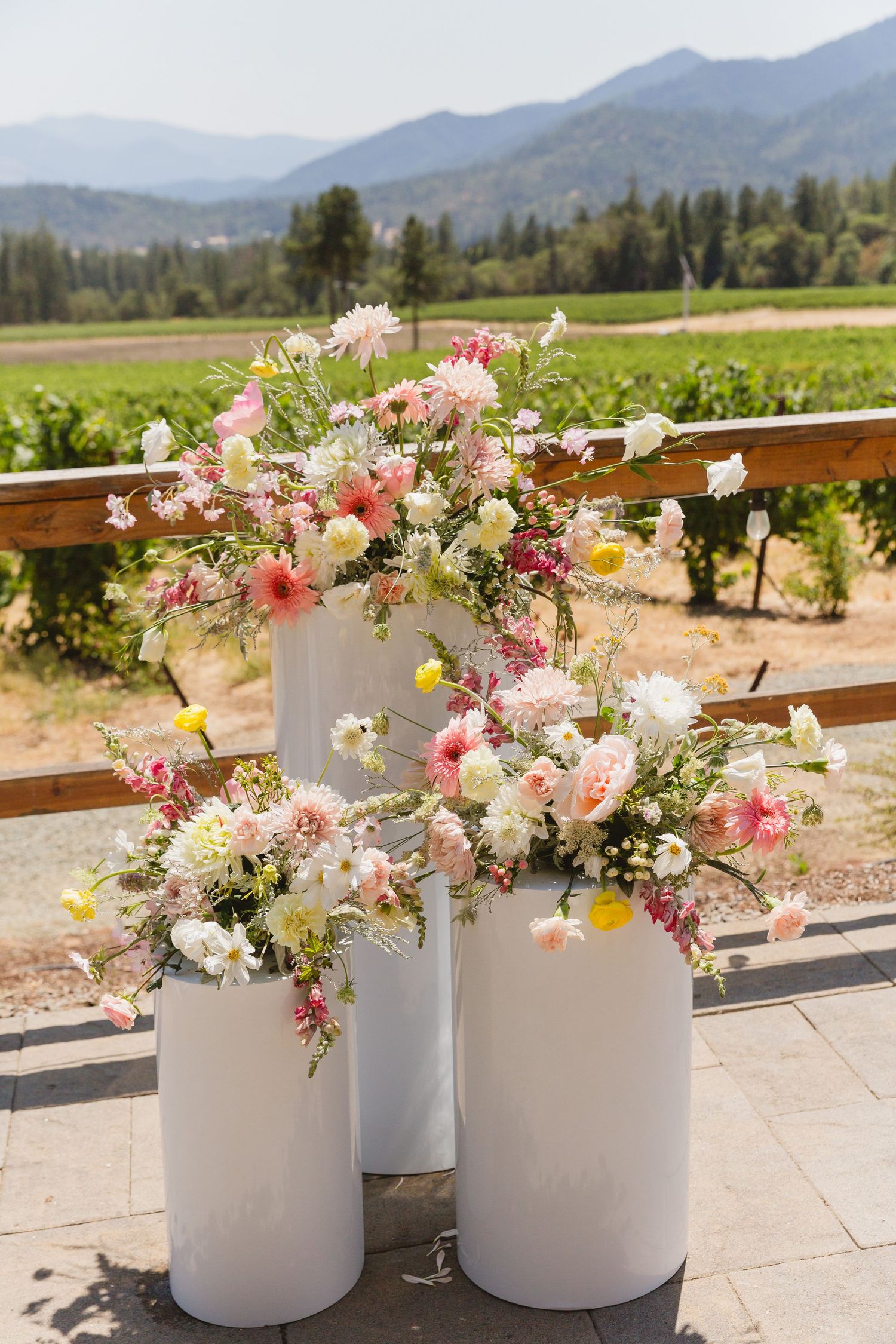 White and pink floral arrangements in ceramic vases decorate a wooden railing with rolling mountains in the background.