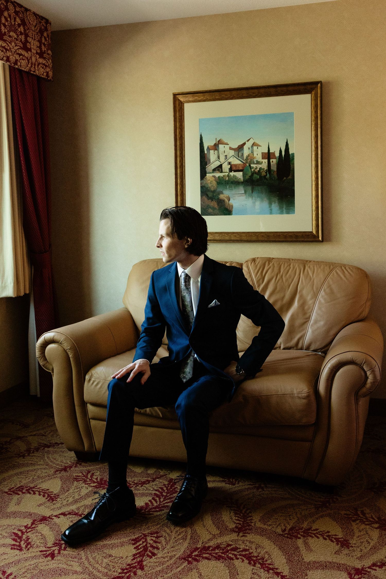 Dapper groom in a navy blue suit sitting on a leather sofa in a hotel room before his wedding.