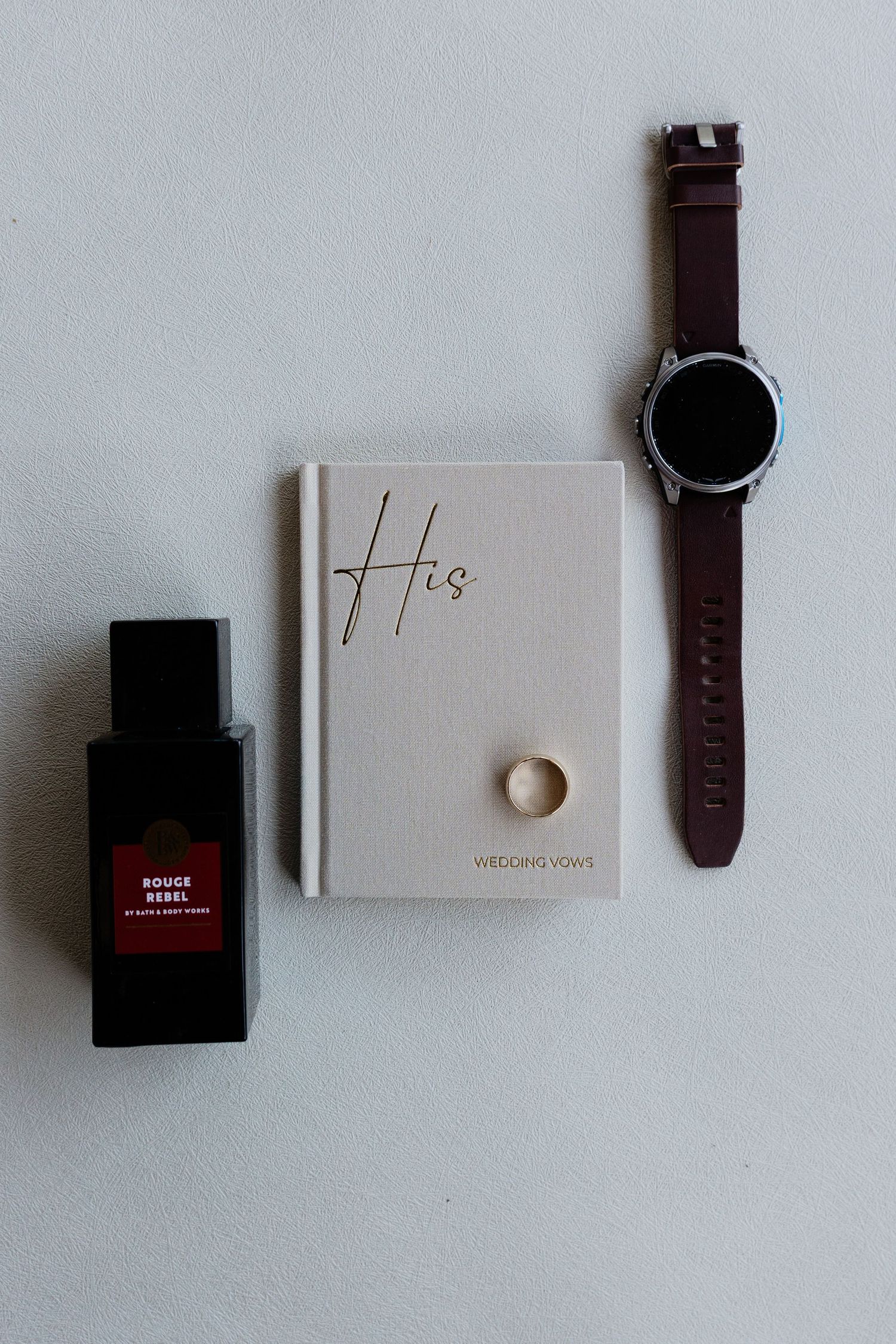 Flat lay of groom wedding essentials including Rouge Rebel cologne, His vows book with ring, and a smartwatch.