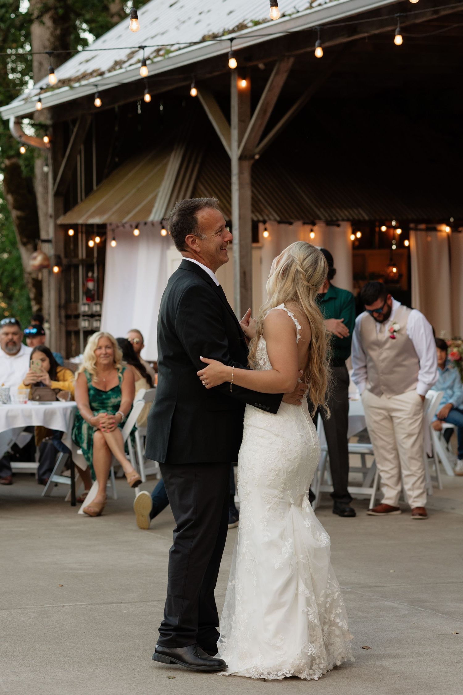 Bride sharing father-daughter dance under string lights at outdoor wedding reception with guests watching.