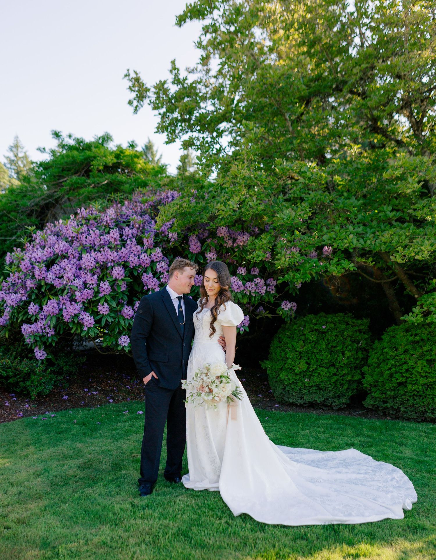 A couple poses in wedding attire in front of blooming purple rhododendrons and lush green trees.