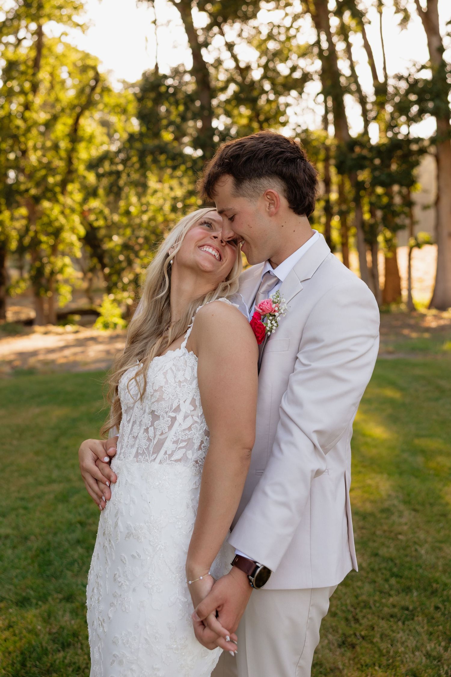 Bride and groom embracing tenderly in golden sunlight surrounded by lush green trees.