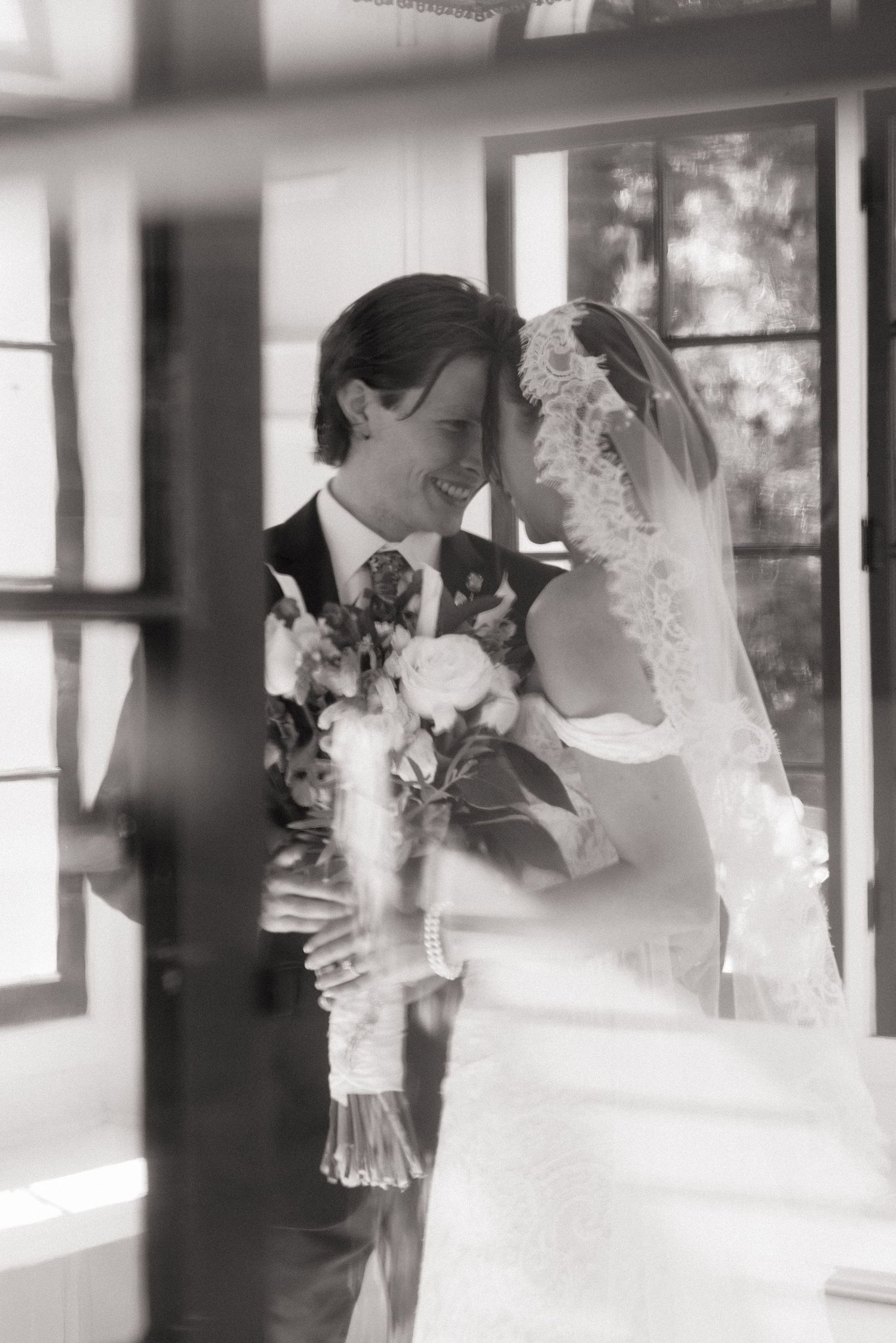 Black and white romantic portrait of bride and groom sharing an intimate moment through a window reflection.