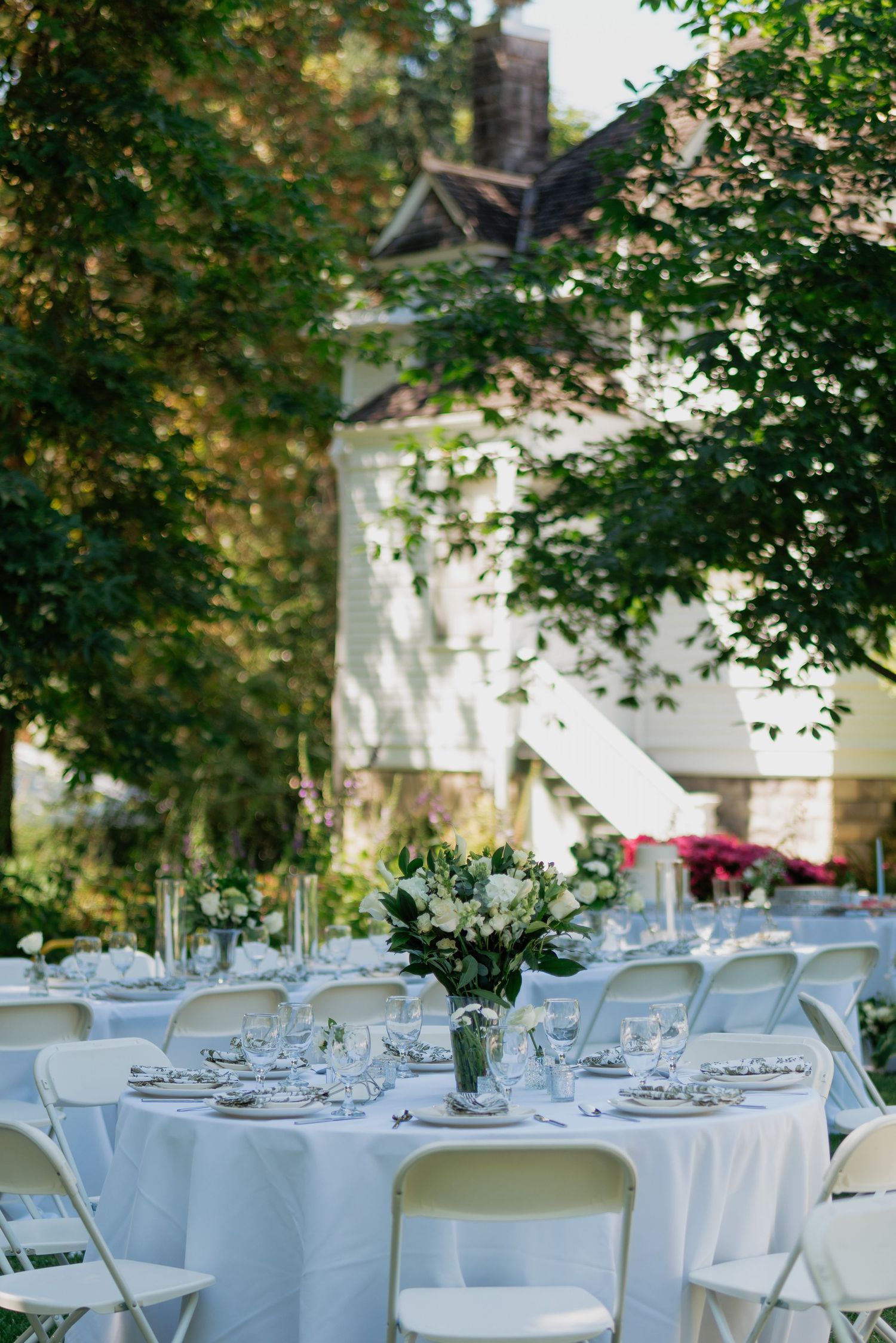 Elegant outdoor wedding reception with white round tables, floral centerpieces, and lush green trees surrounding a white venue.