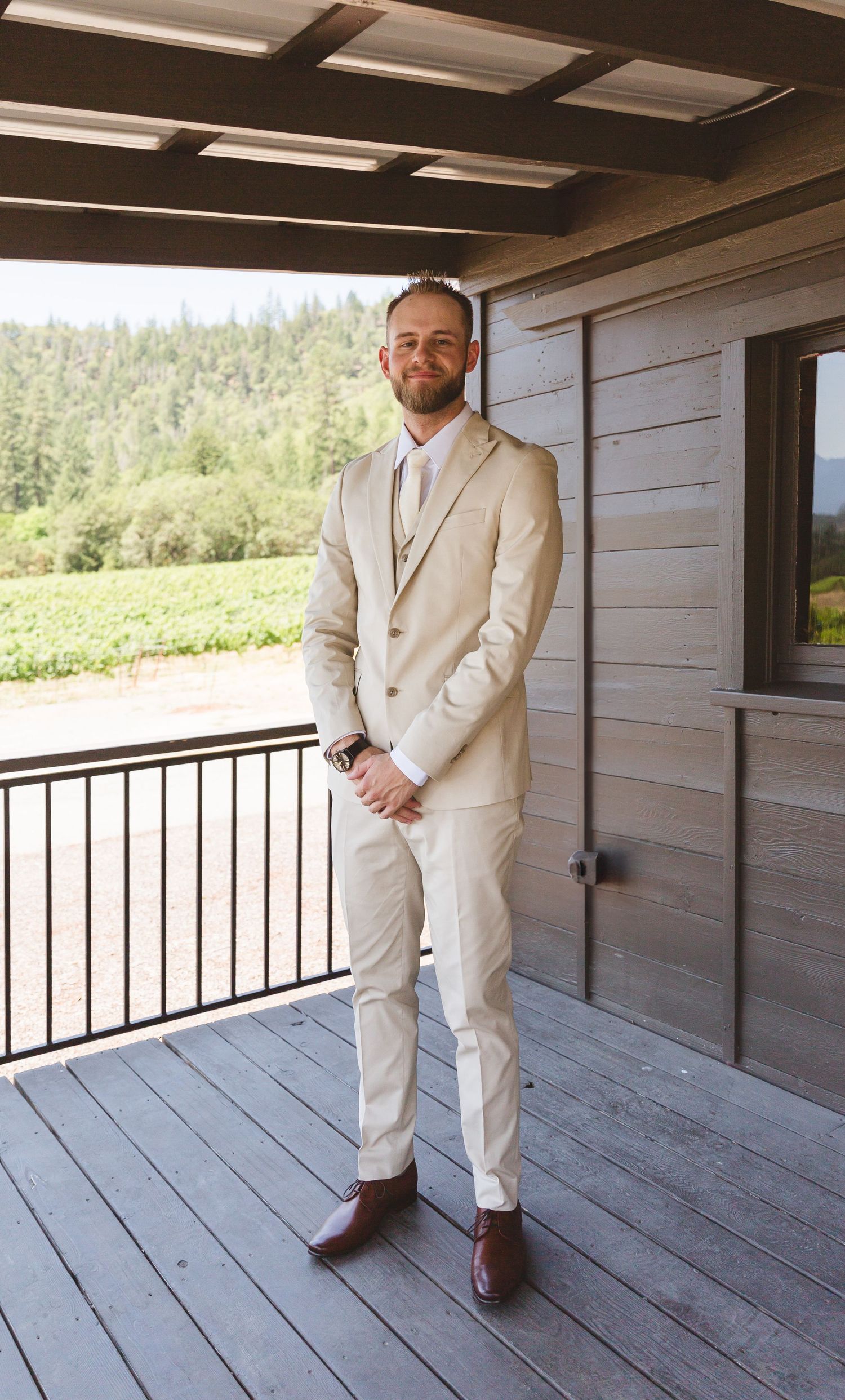 A groom wearing a light beige suit stands on a wooden deck with mountain views in the background.