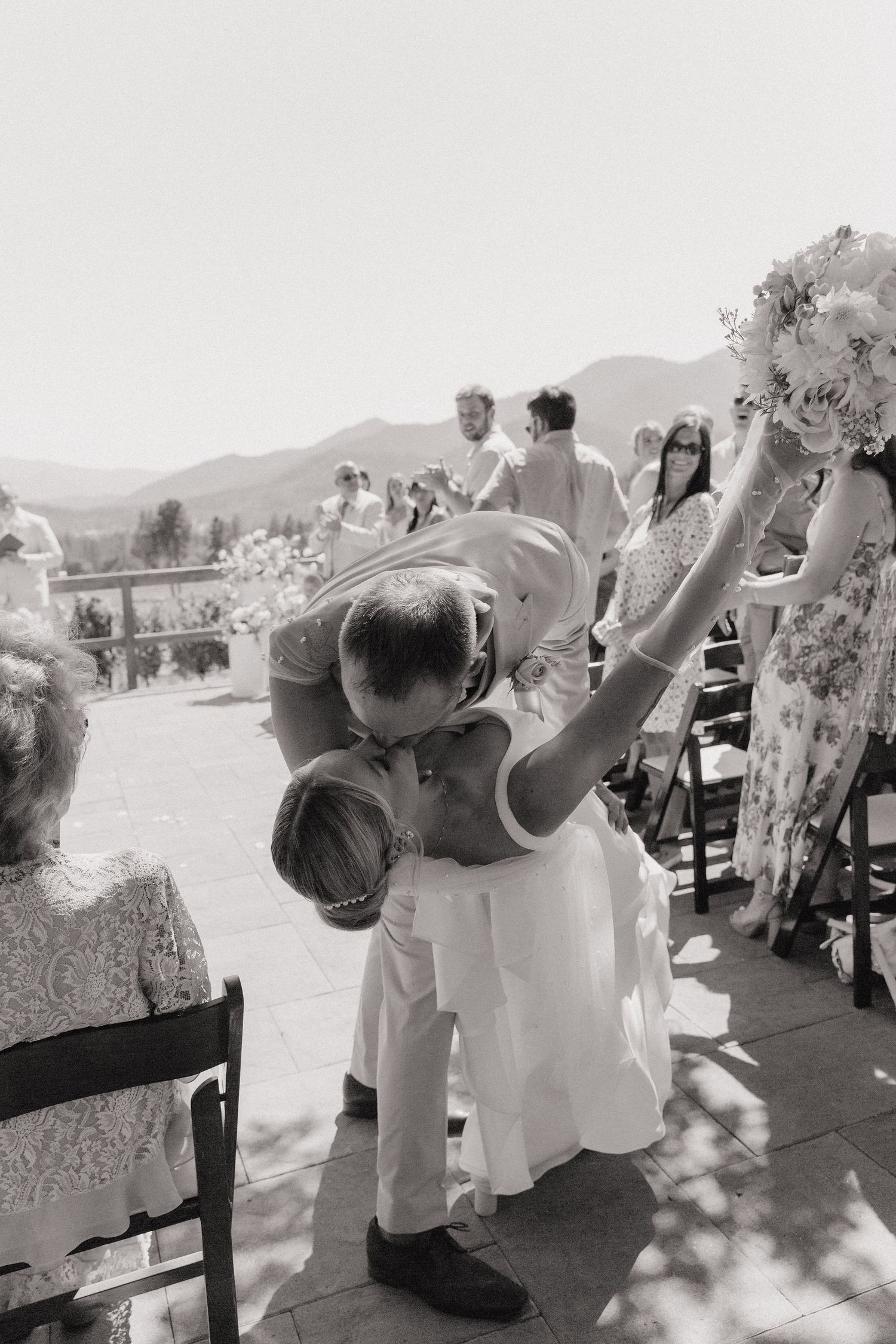Black and white photo of newlyweds sharing a kiss at outdoor wedding ceremony with mountain views.