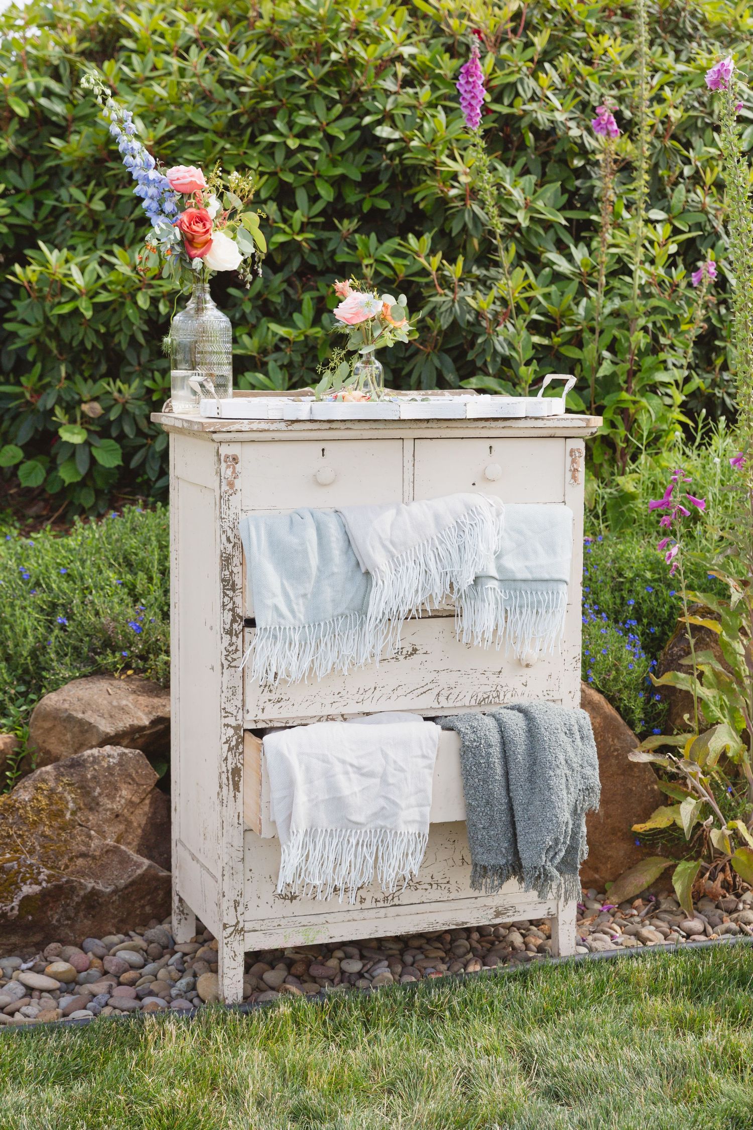 Vintage white distressed sideboard decorated with draped blankets and floral arrangements sits in a garden setting.