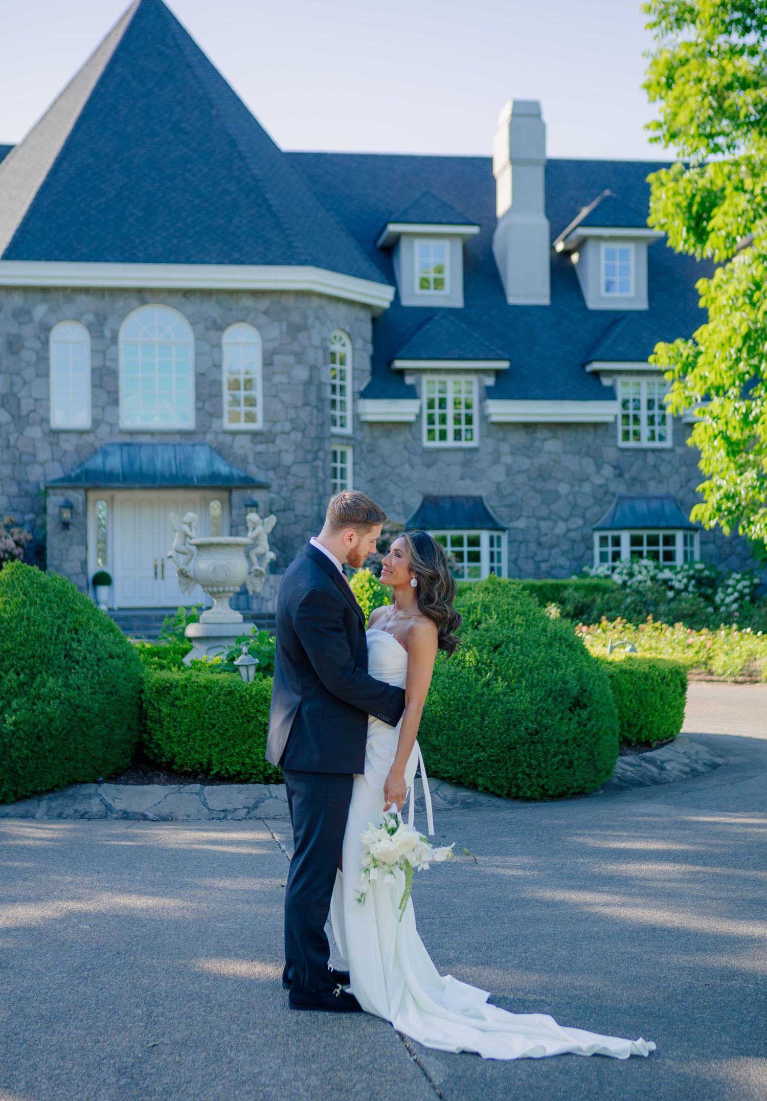 A couple embraces in front of an elegant stone mansion with manicured hedges and a dark blue roof on a sunny day.