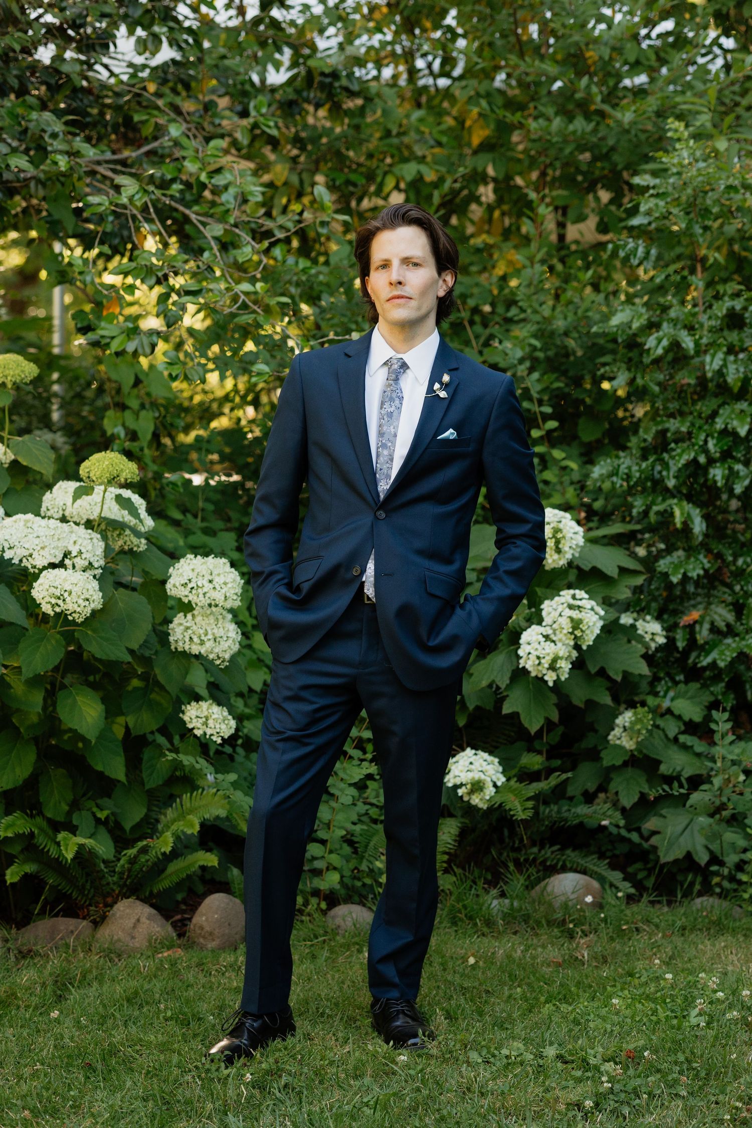 Handsome groom in a navy suit with floral tie standing in a garden surrounded by white hydrangeas.