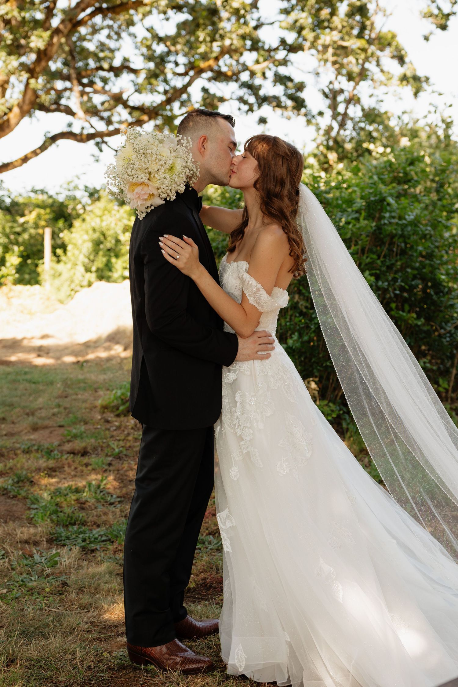 Bride and groom sharing a romantic kiss outdoors, bride holding white floral bouquet with long veil flowing.