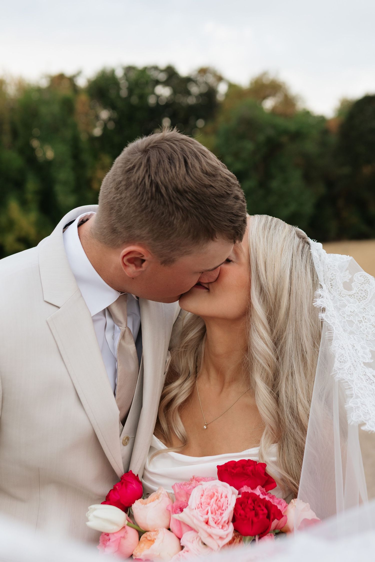 A bride and groom share a romantic kiss, she holds a bouquet of pink and red roses, wearing a veil and white gown.