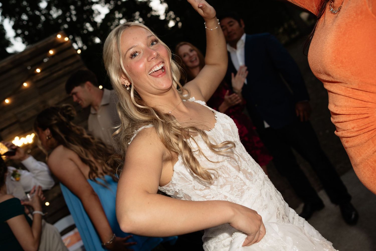Joyful blonde bride in lace gown laughing and dancing at outdoor wedding reception celebration.