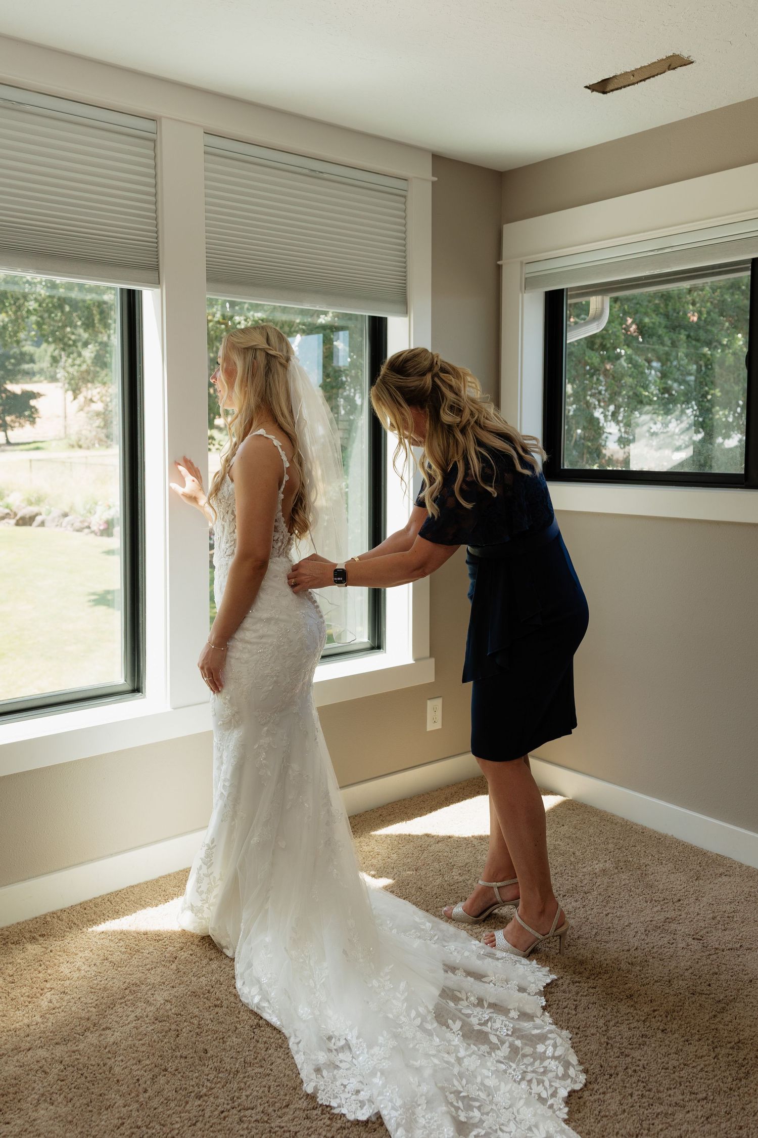 Mother helping bride fasten her lace wedding gown before the ceremony in a sunlit room.