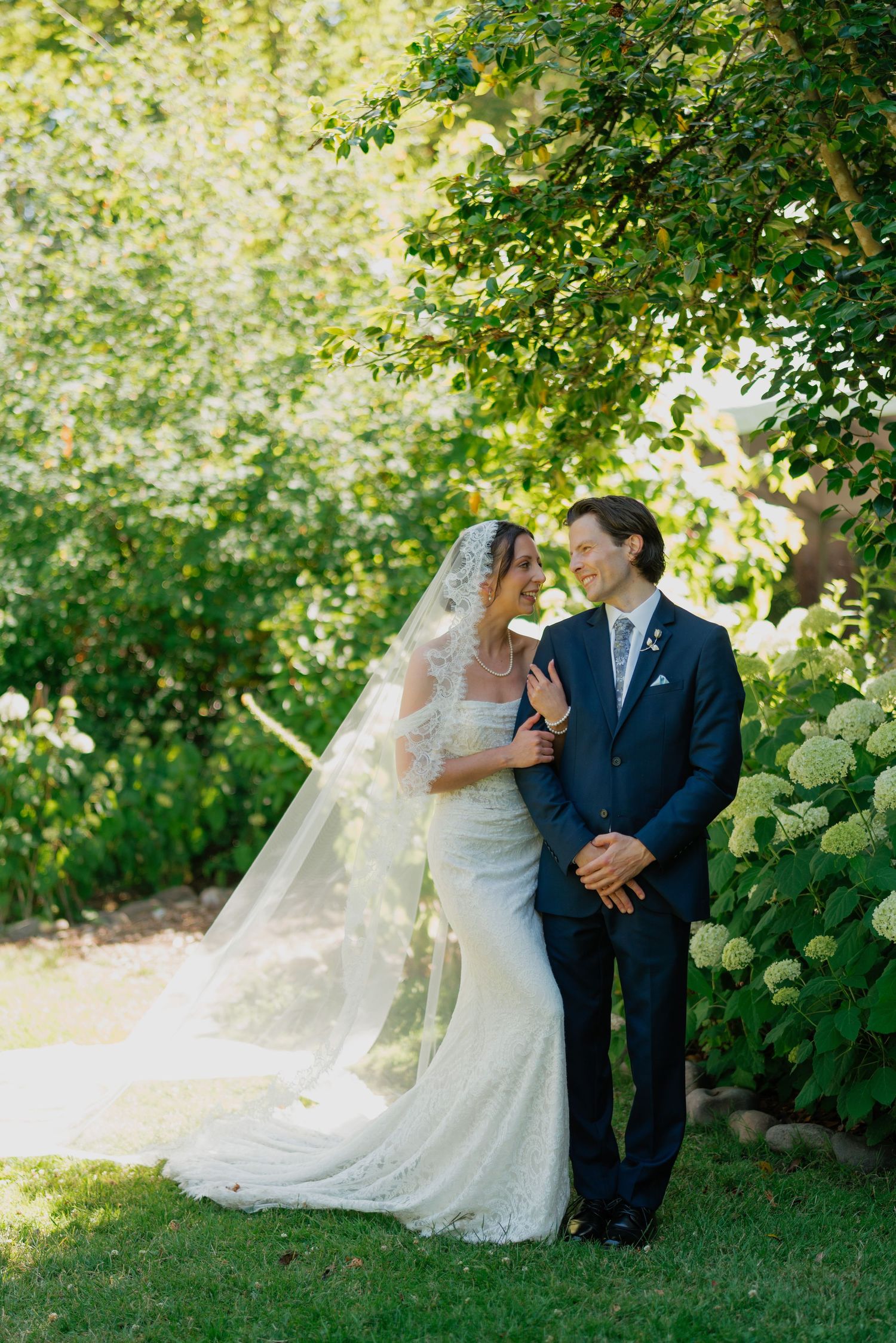 Bride in lace wedding gown with cathedral veil and groom in navy suit share romantic moment in sunlit garden.