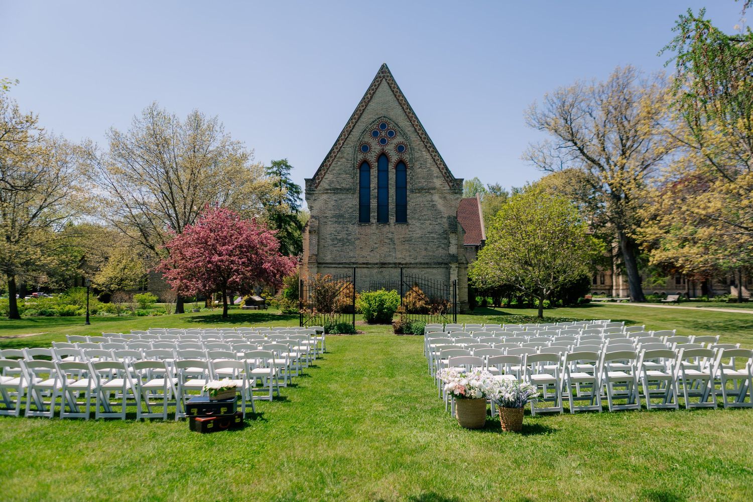 White wedding chairs line up towards a rustic chapel on a scenic green lawn surrounded by spring blooming trees.