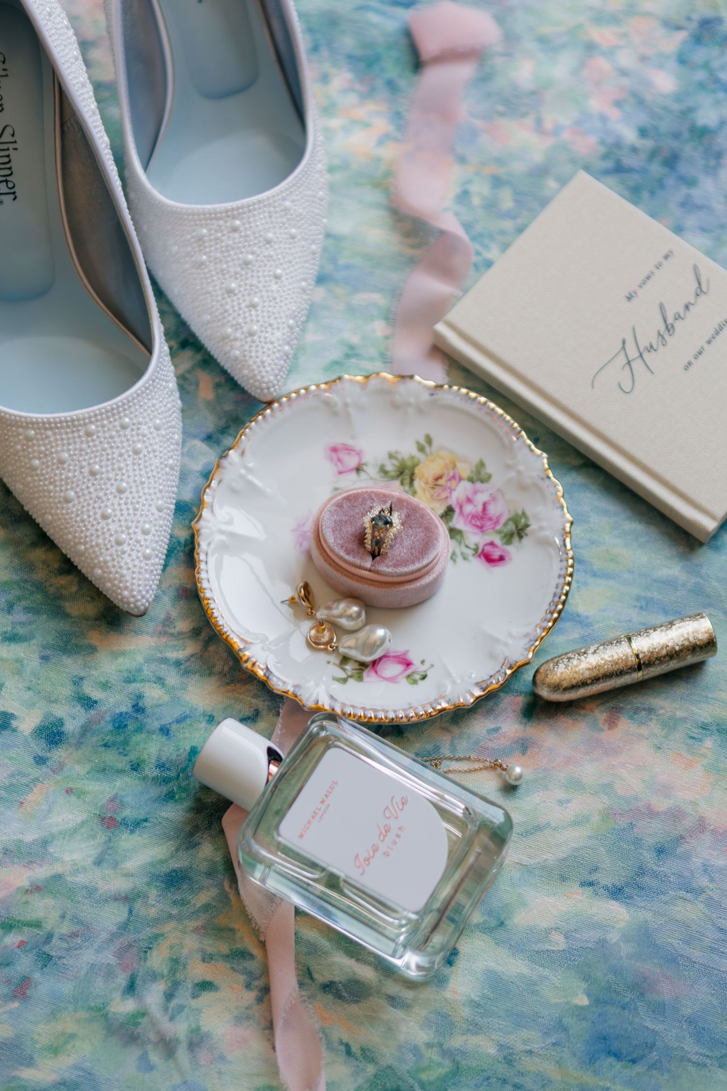 Wedding accessories including sparkly white heels, floral china plate, perfume bottle, and pink velvet ring box on blue surface.