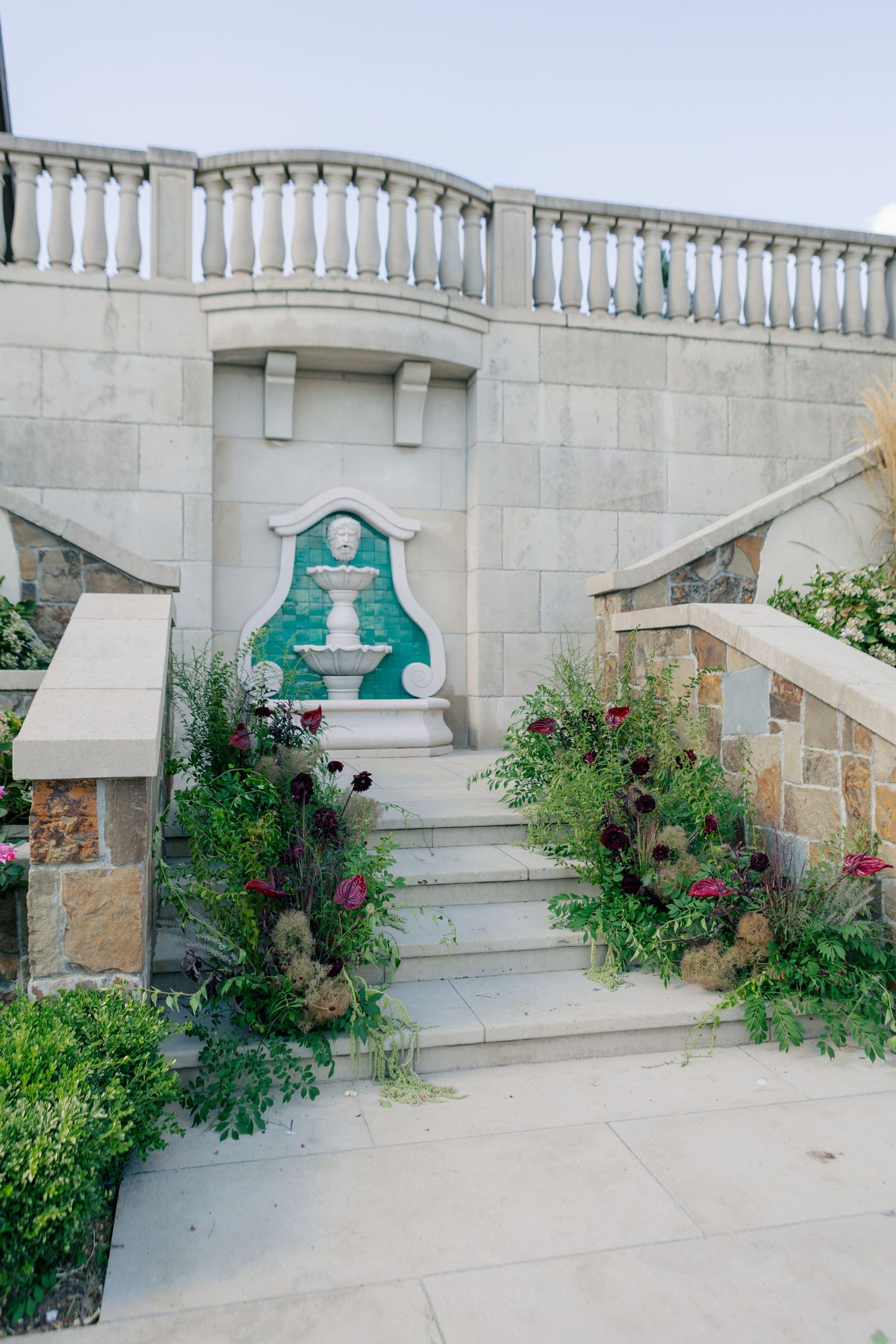 Stone steps with ornate balustrade lead to an elevated balcony surrounded by colorful flowering plants and landscaping elements.