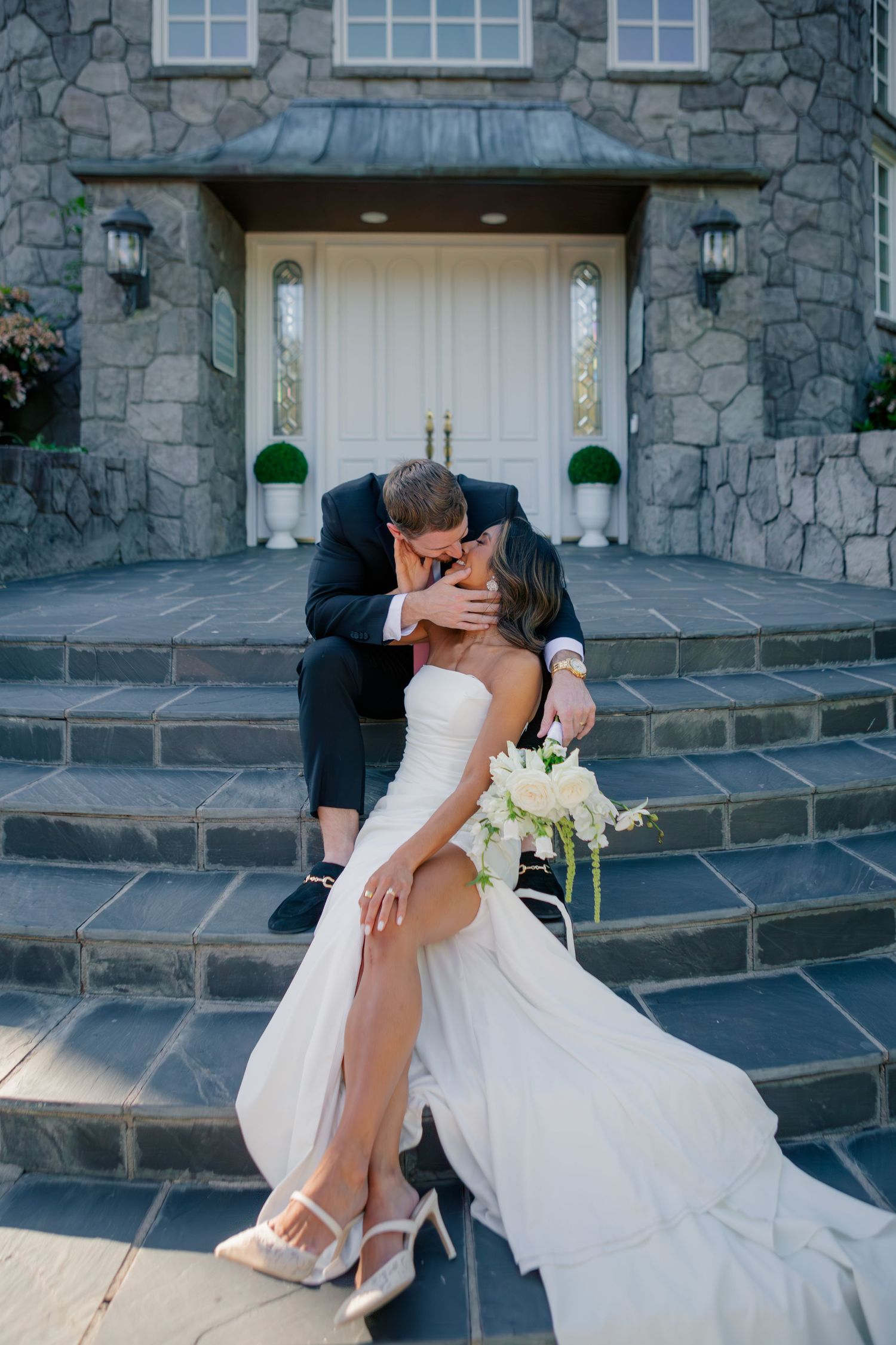 A romantic wedding couple kiss on stone steps in front of an elegant stone building with white door.