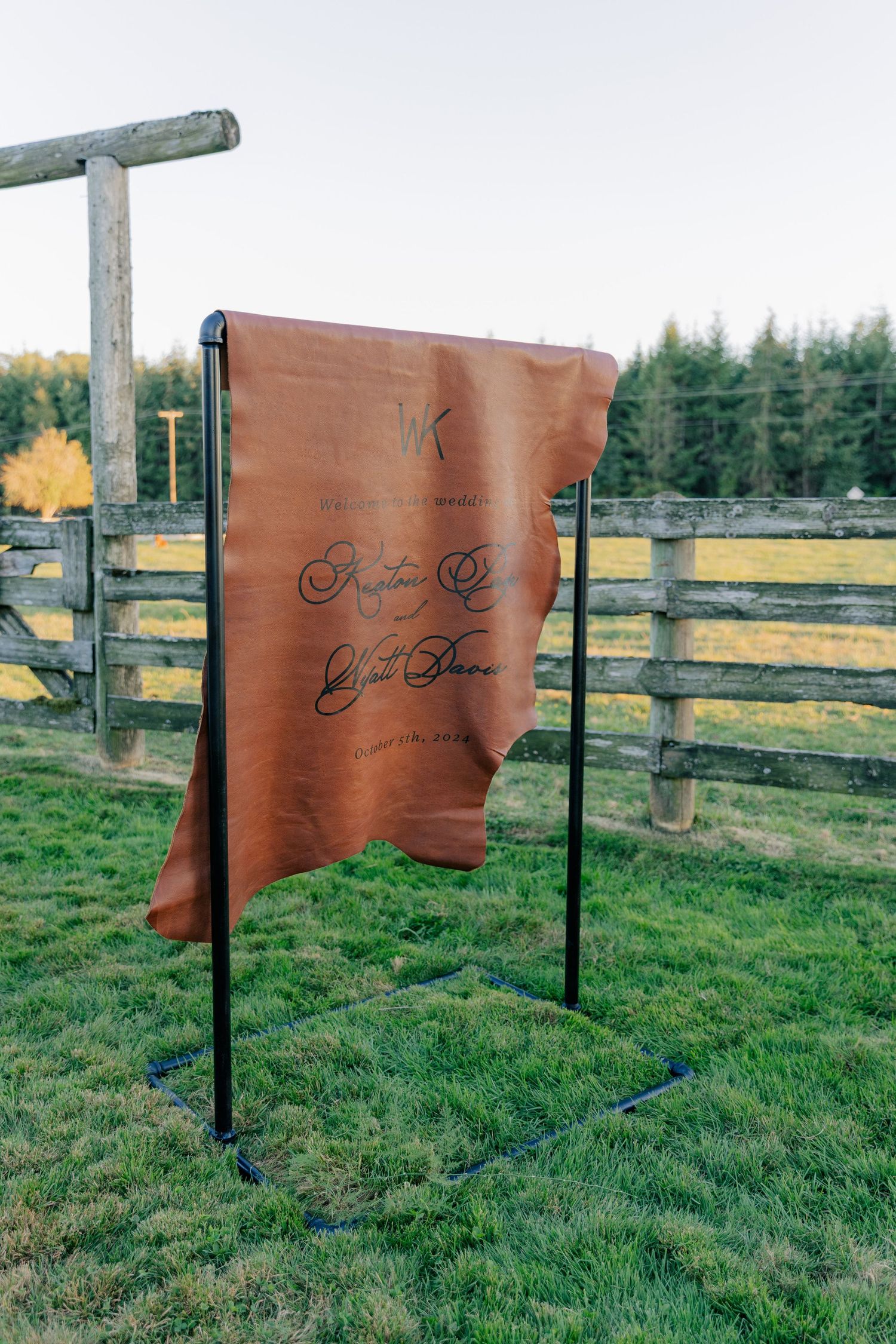 A leather banner with text, fluttering in the wind, hangs between wooden posts on grassy farmland with a treeline behind it.