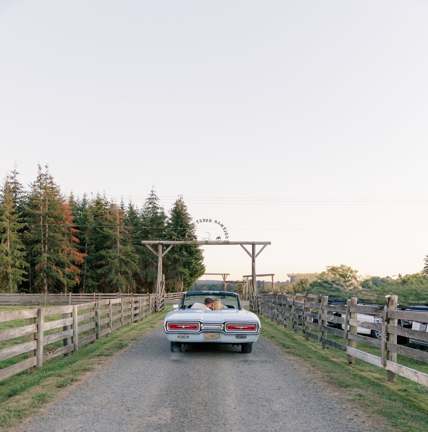 A vintage convertible car drives along a dirt road between wooden fences with evergreen trees in the background.
