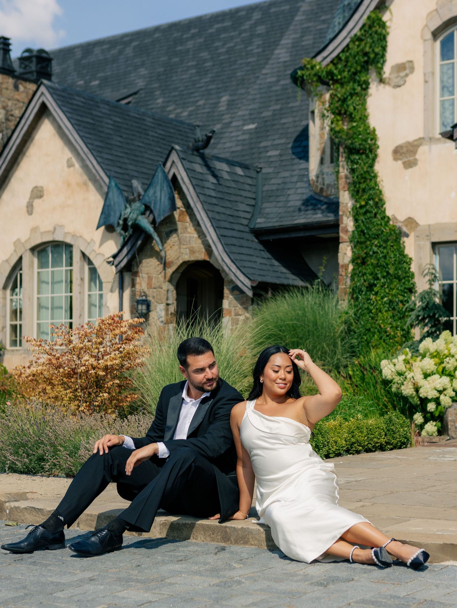 A couple lounging casually on outdoor stone steps against a luxurious estate backdrop in elegant wedding attire.