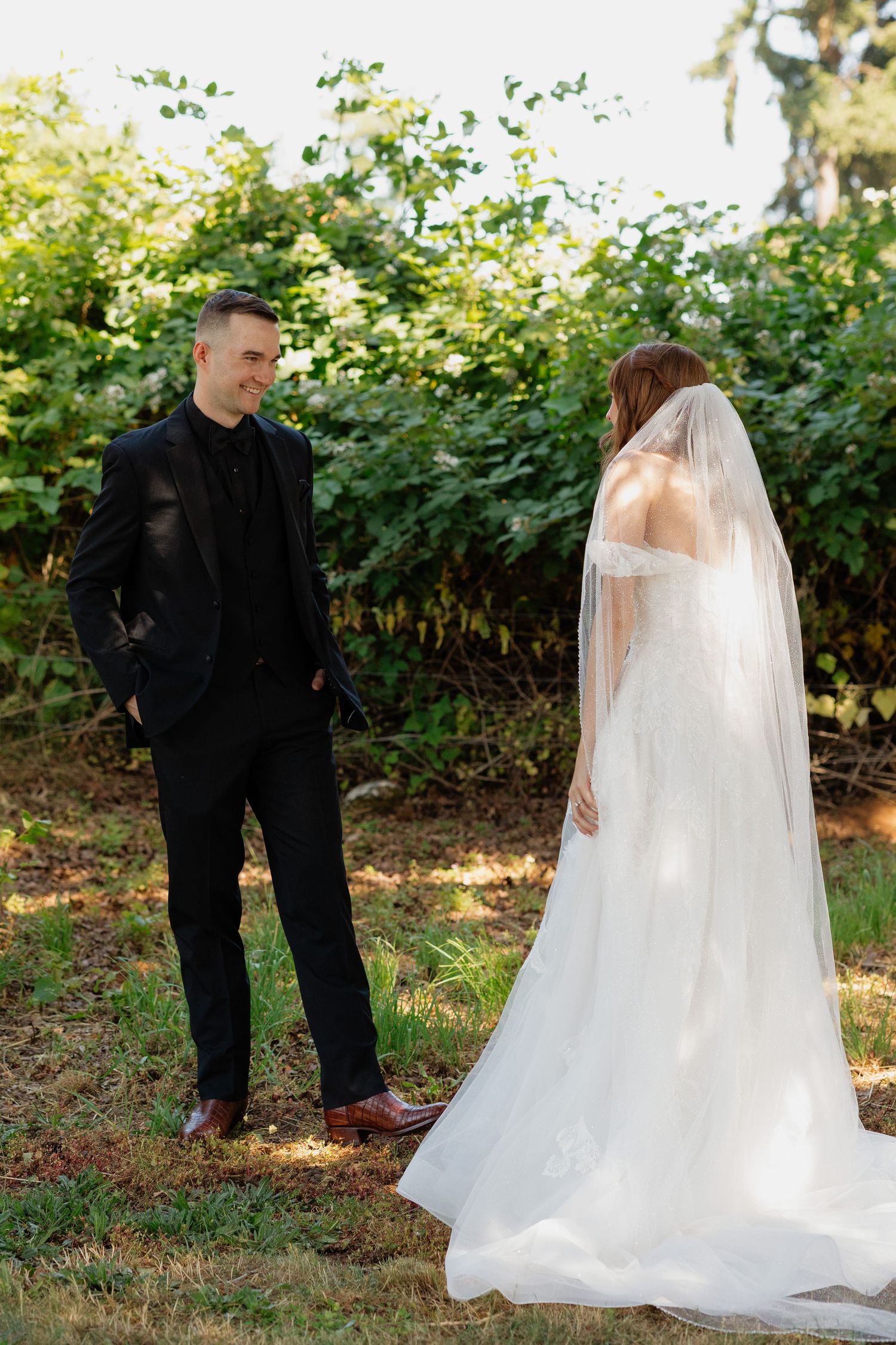 Smiling groom in black suit faces bride in white wedding gown with long veil in lush garden setting.