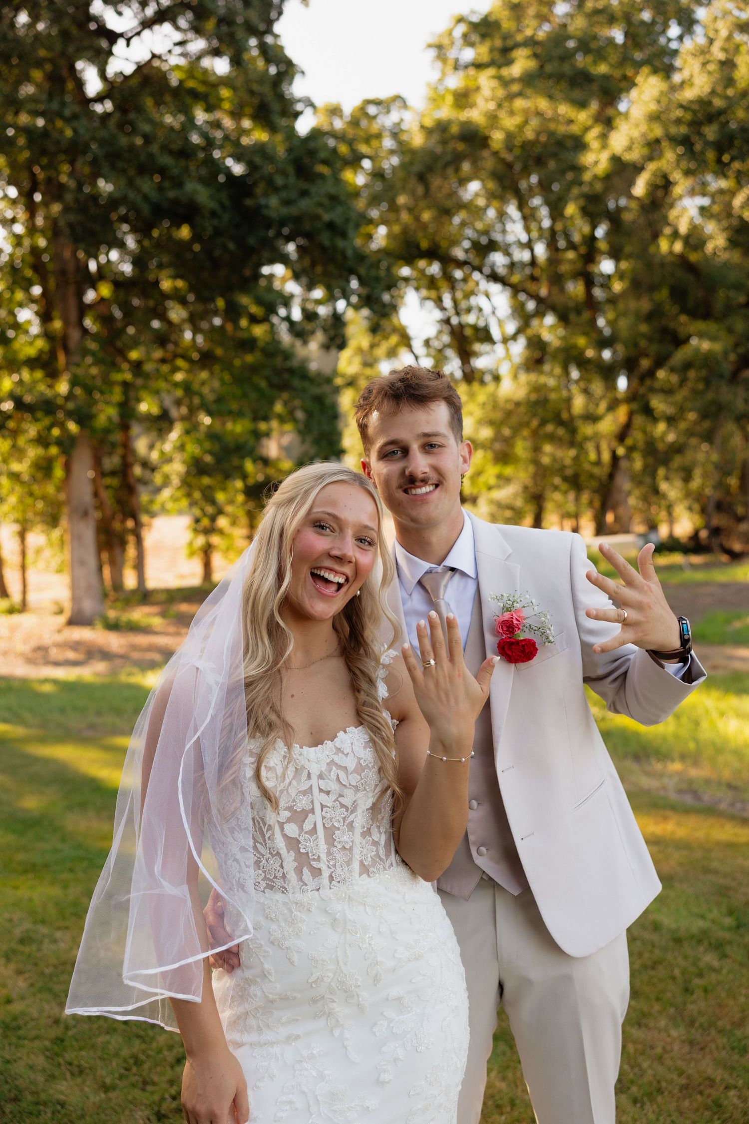 Happy newlyweds showing off wedding rings while smiling outdoors in golden hour light after ceremony.