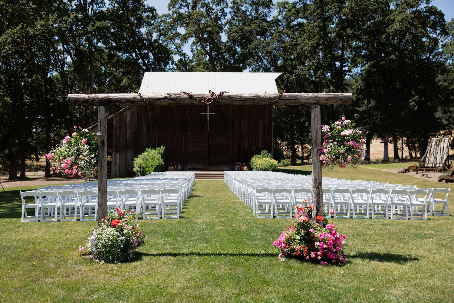 Outdoor wedding ceremony setup with white chairs, floral arrangements, rustic barn backdrop, and wooden arbor on a sunny day.
