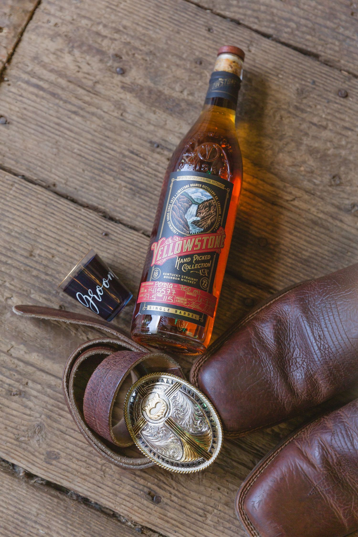 A bottle of Jagermeister liquor next to brown leather boots and a decorative belt buckle on wooden floorboards.