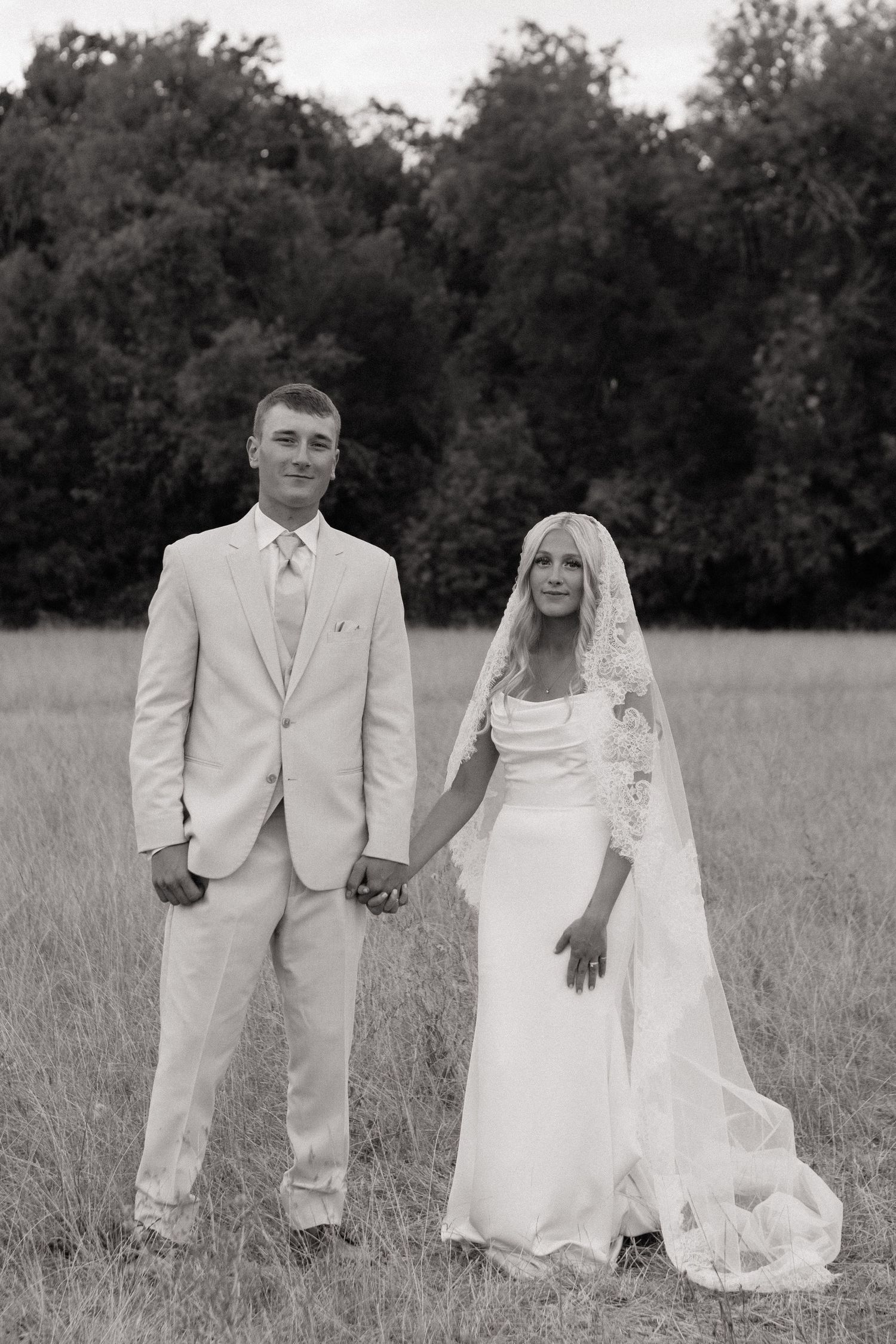 Black and white photo of a bride and groom holding hands in a field, wearing wedding attire with trees in background.