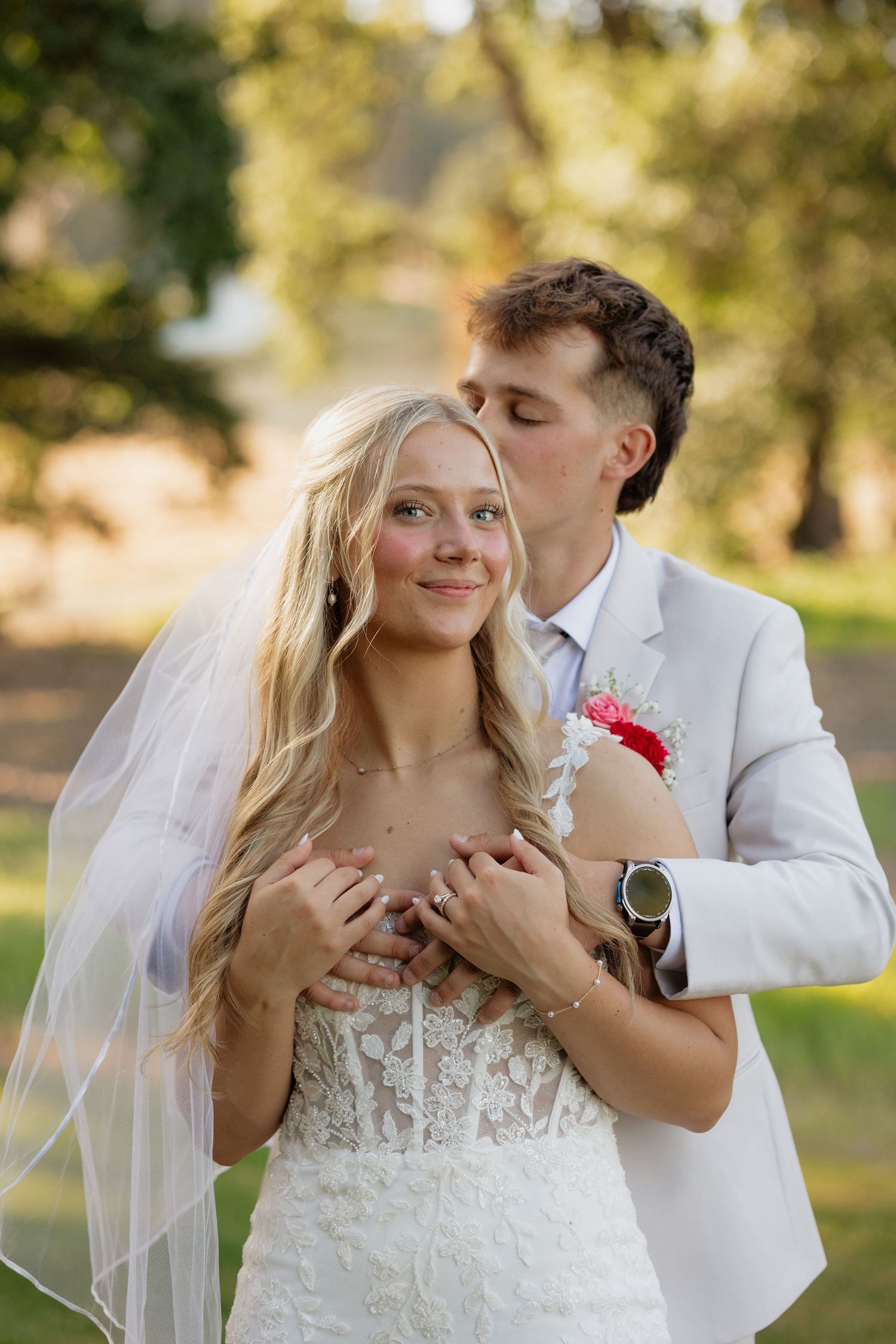 Groom in cream suit kissing bride on cheek while embracing her from behind in sunny garden.