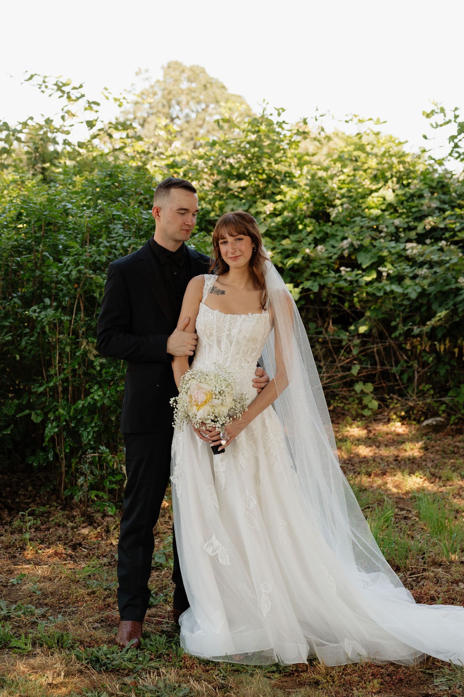 Bride in white lace gown with veil and groom in black suit posing outdoors near green foliage.