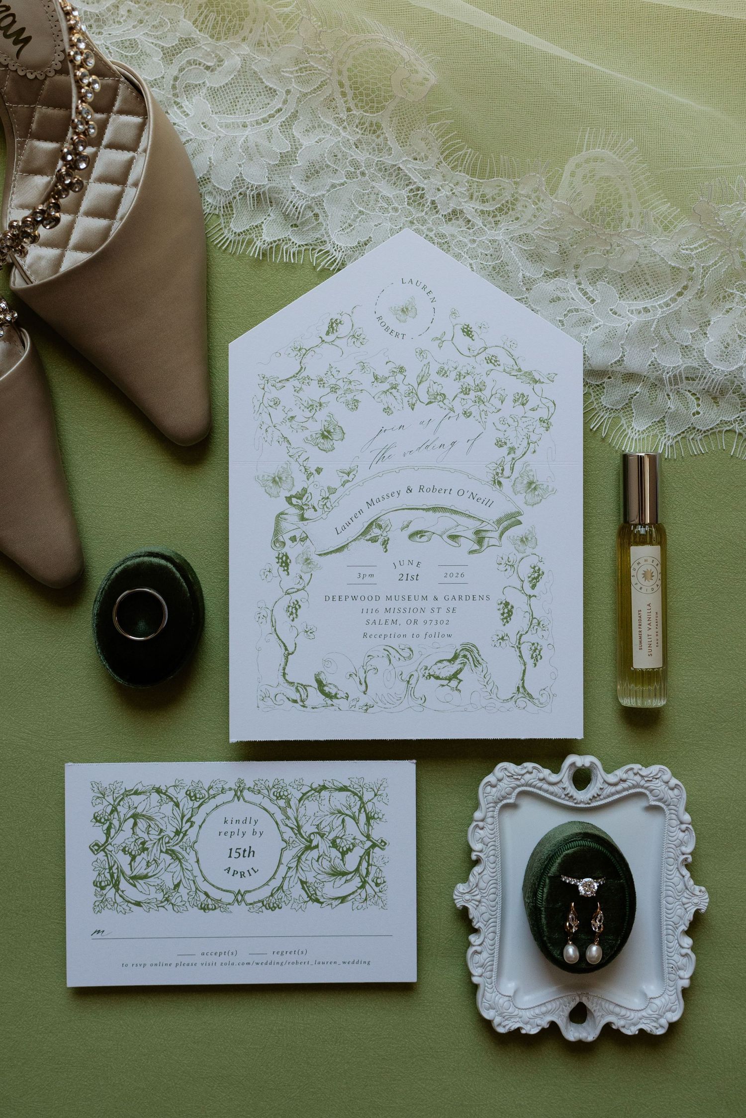 Elegant green and white wedding invitation suite flat lay with earrings, perfume, rings, and bridal veil on green background.